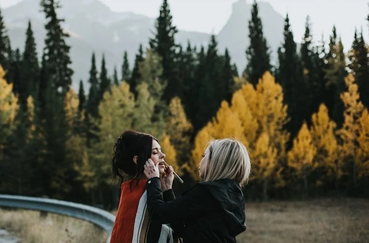 Two women outdoors, one applying makeup to the other, with a backdrop of a forest of evergreen and yellow-leaved trees.