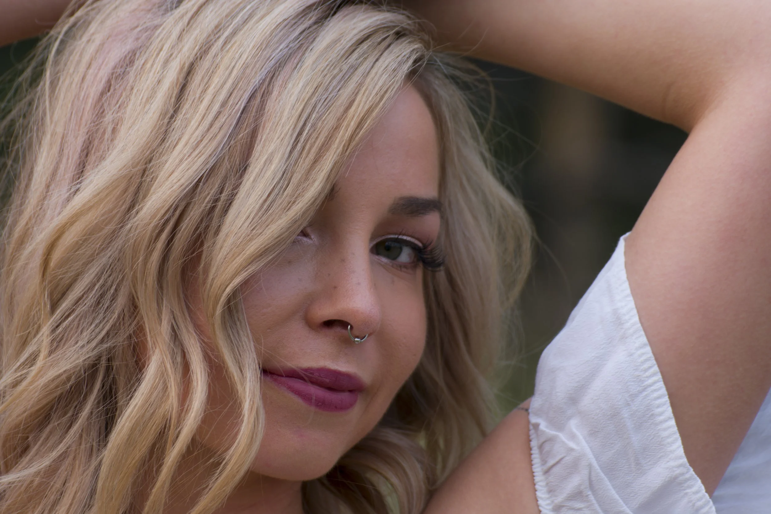 A woman with long blonde hair, blue eyes, and a septum piercing, looking at the camera with her arm raised.