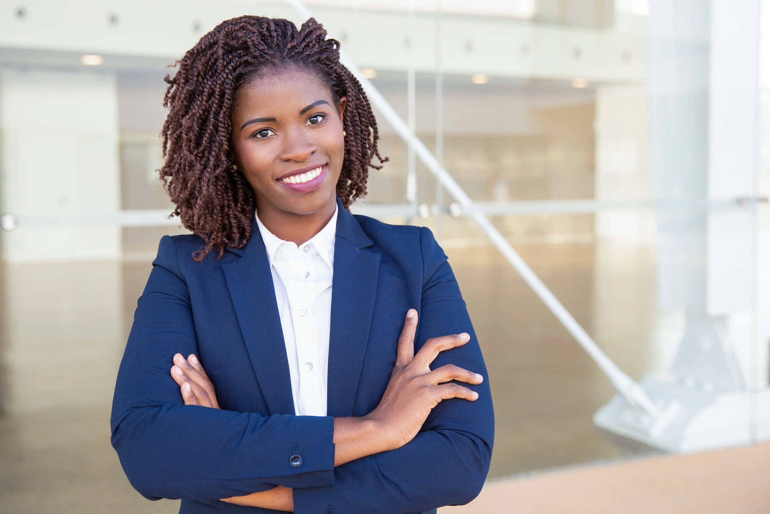 A professional woman with curly hair, wearing a navy blazer and white shirt, smiling with arms crossed in an office setting.