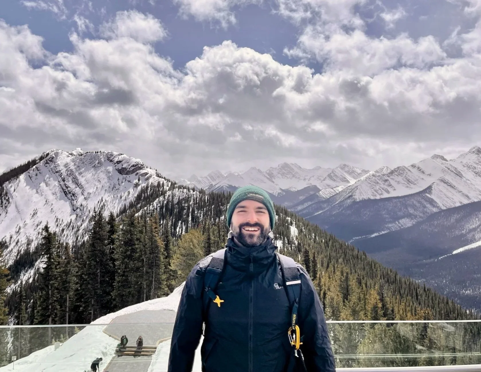 A man smiling in winter mountain landscape with snow-covered peaks, evergreen trees, and partly cloudy sky.