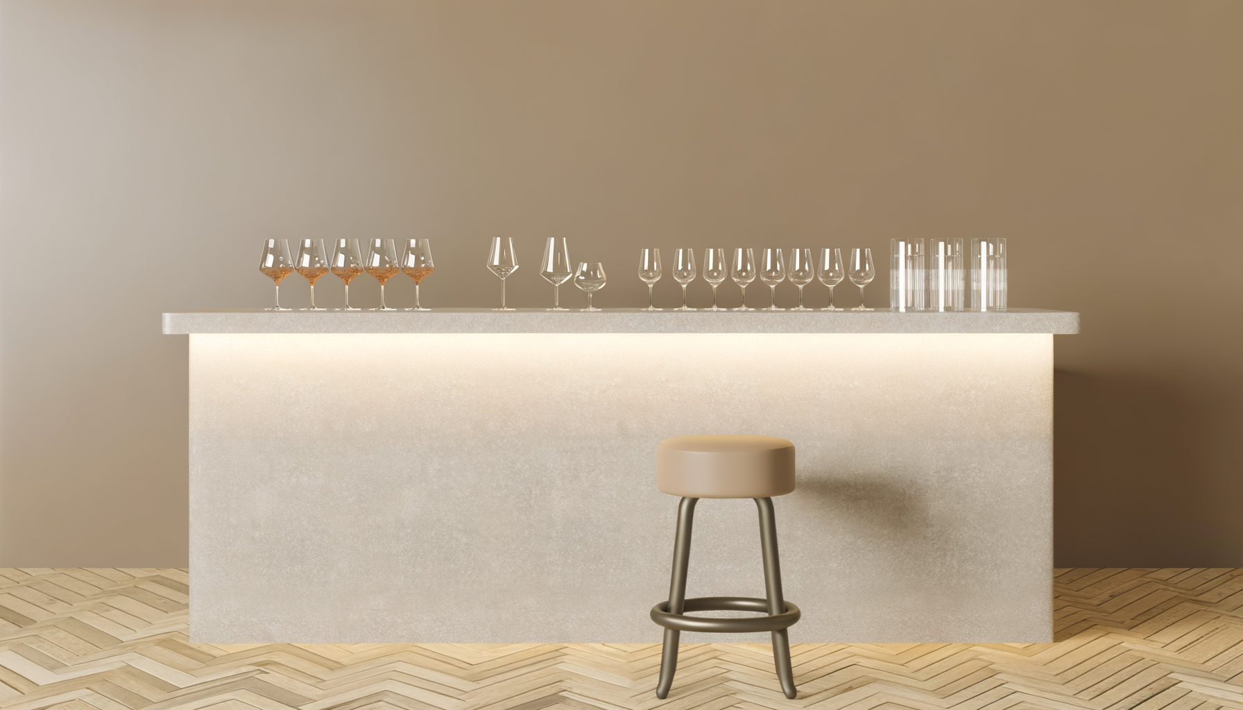 A modern bar with a beige bar counter, various empty wine glasses and shot glasses on top, a beige stool with a round seat in front, and a wooden herringbone floor.