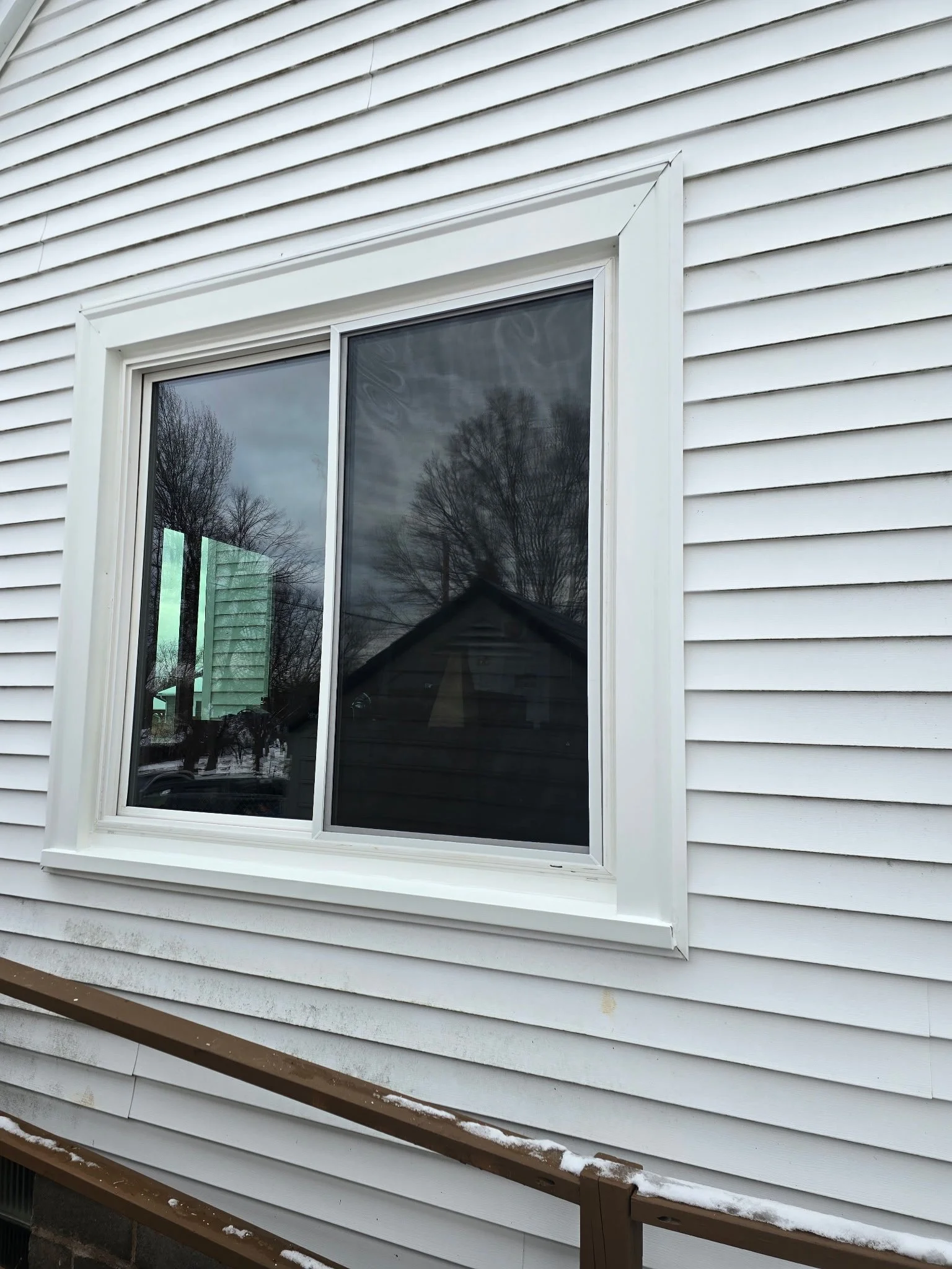 Exterior view of a house with white vinyl siding and a window with white trim. Reflection of trees and a cloudy sky in the window and a neighboring house visible.