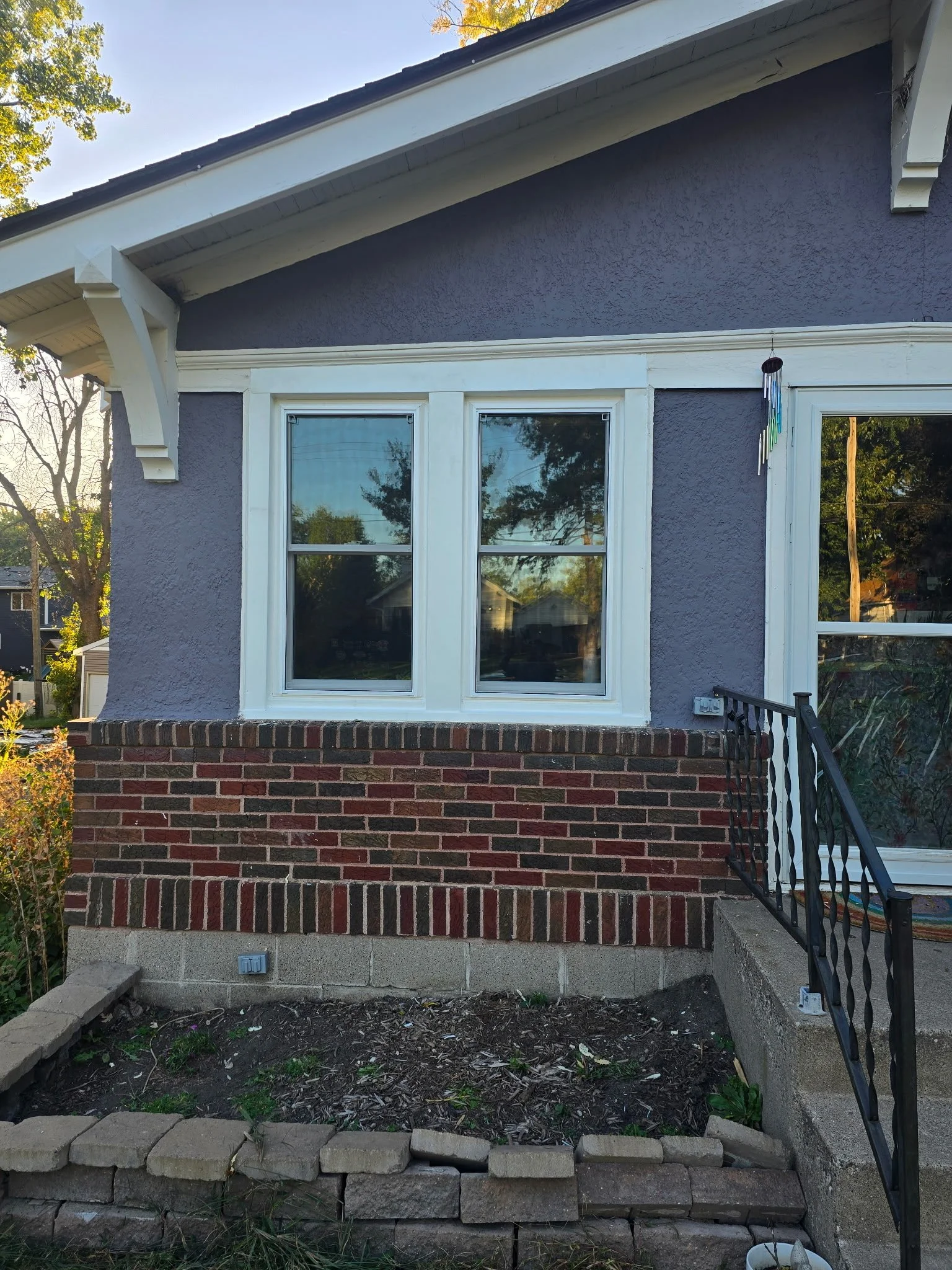 The front of a house with a purple exterior, white trim, two large windows, and a brick foundation. There is a small garden bed with stones in the foreground and a black railing on the right side. A wind chime hangs near the window entrance.