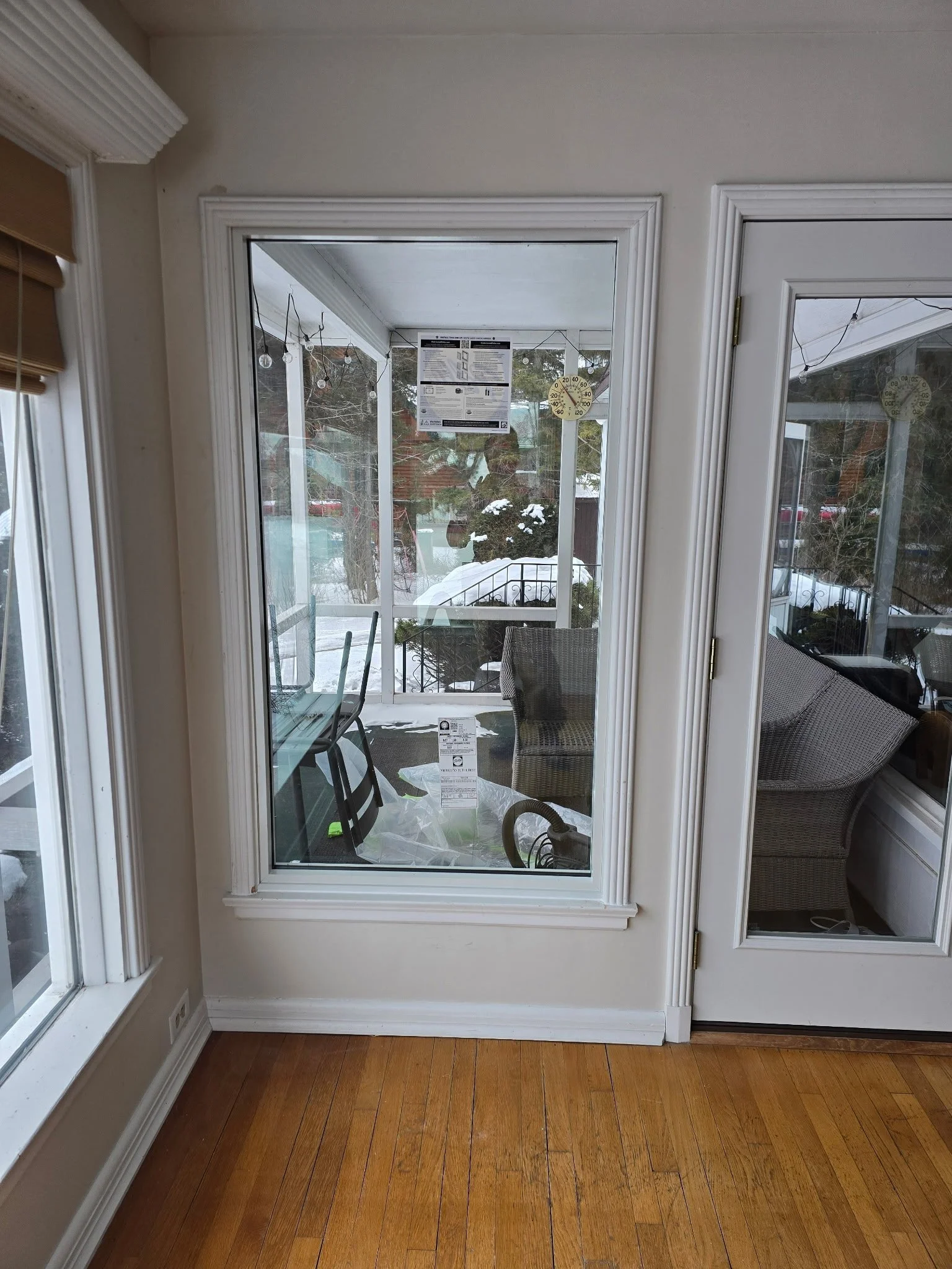 View through glass door and windows showing a porch with chairs and table, snow on the ground and trees outside.