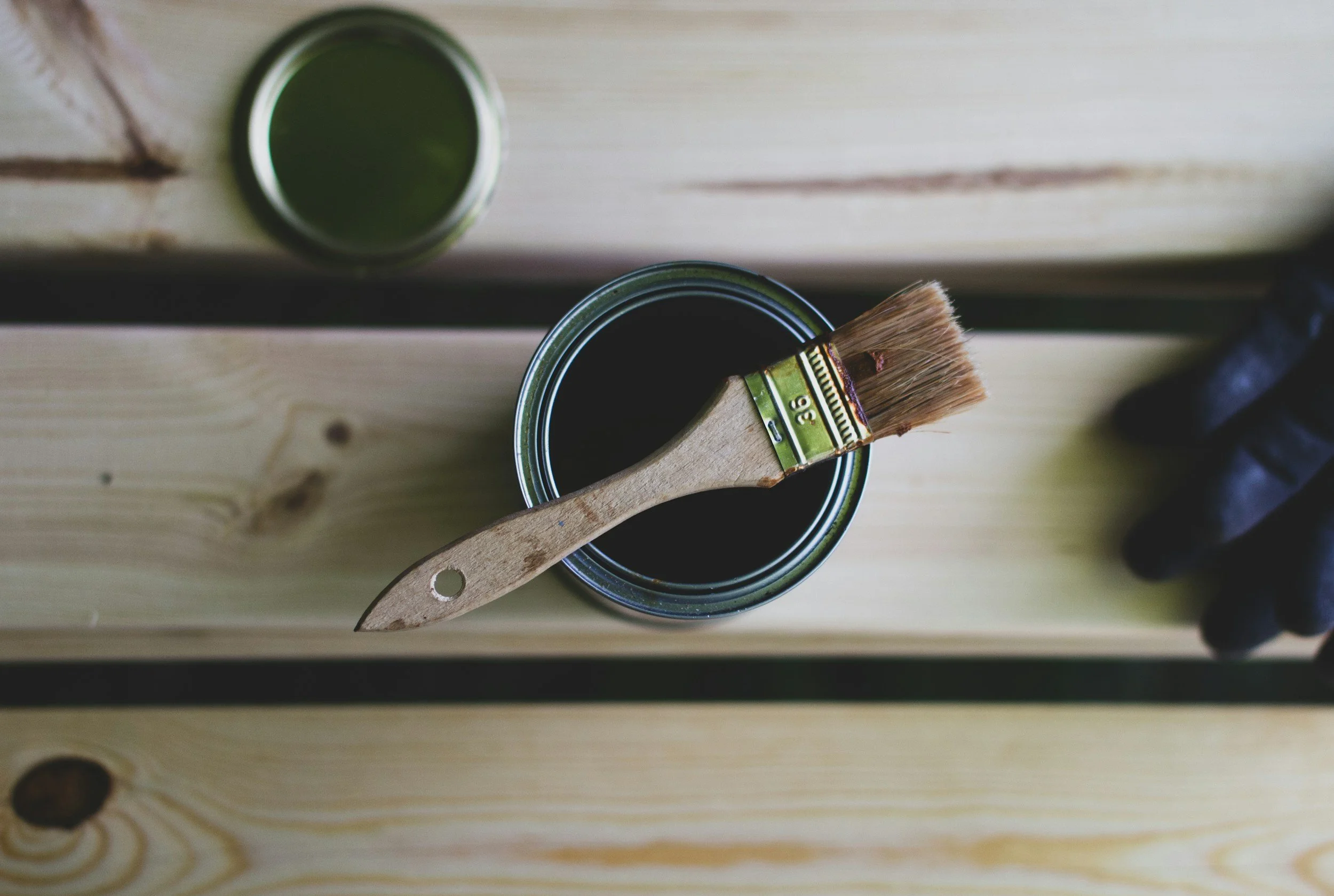 A wooden tabletop with a can of black paint, a paintbrush resting on the rim, and a small container of green paint in the background.