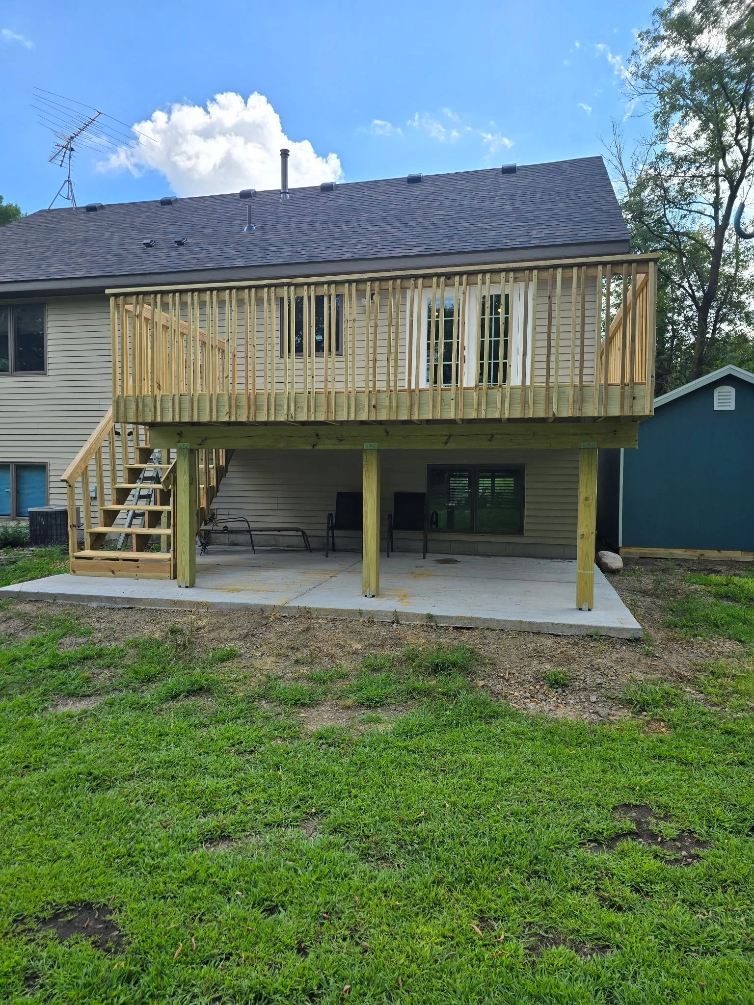 Back of a two-story house with a newly built wooden deck on the upper level. The deck has a railing, stairs, and a door leading inside. There is a concrete patio below with some outdoor furniture. The house has beige siding, and part of another house