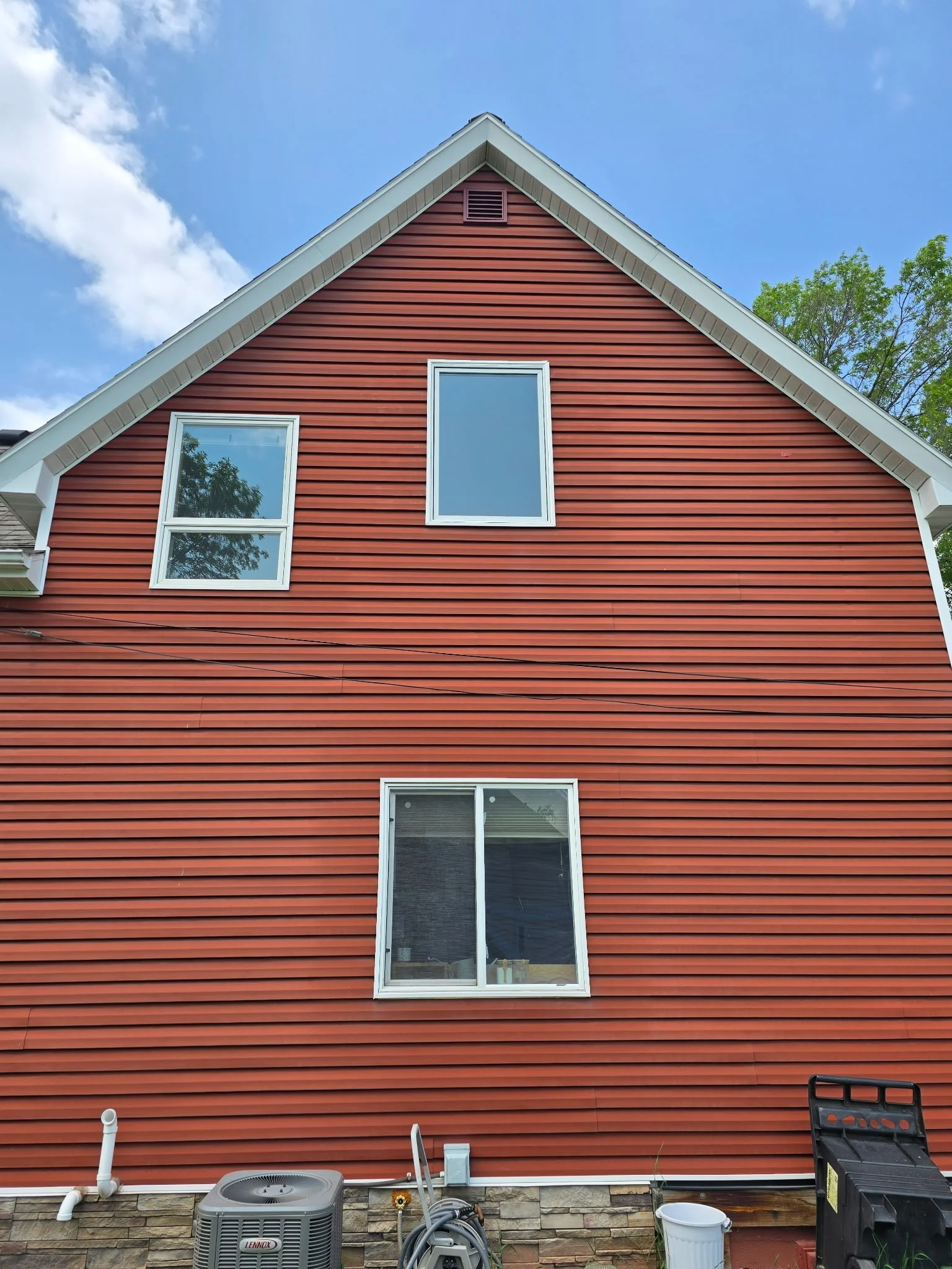 The exterior of a red house with white-trimmed windows, a stone foundation, and an air conditioning unit at the base, under a blue sky with some clouds and green trees in the background.