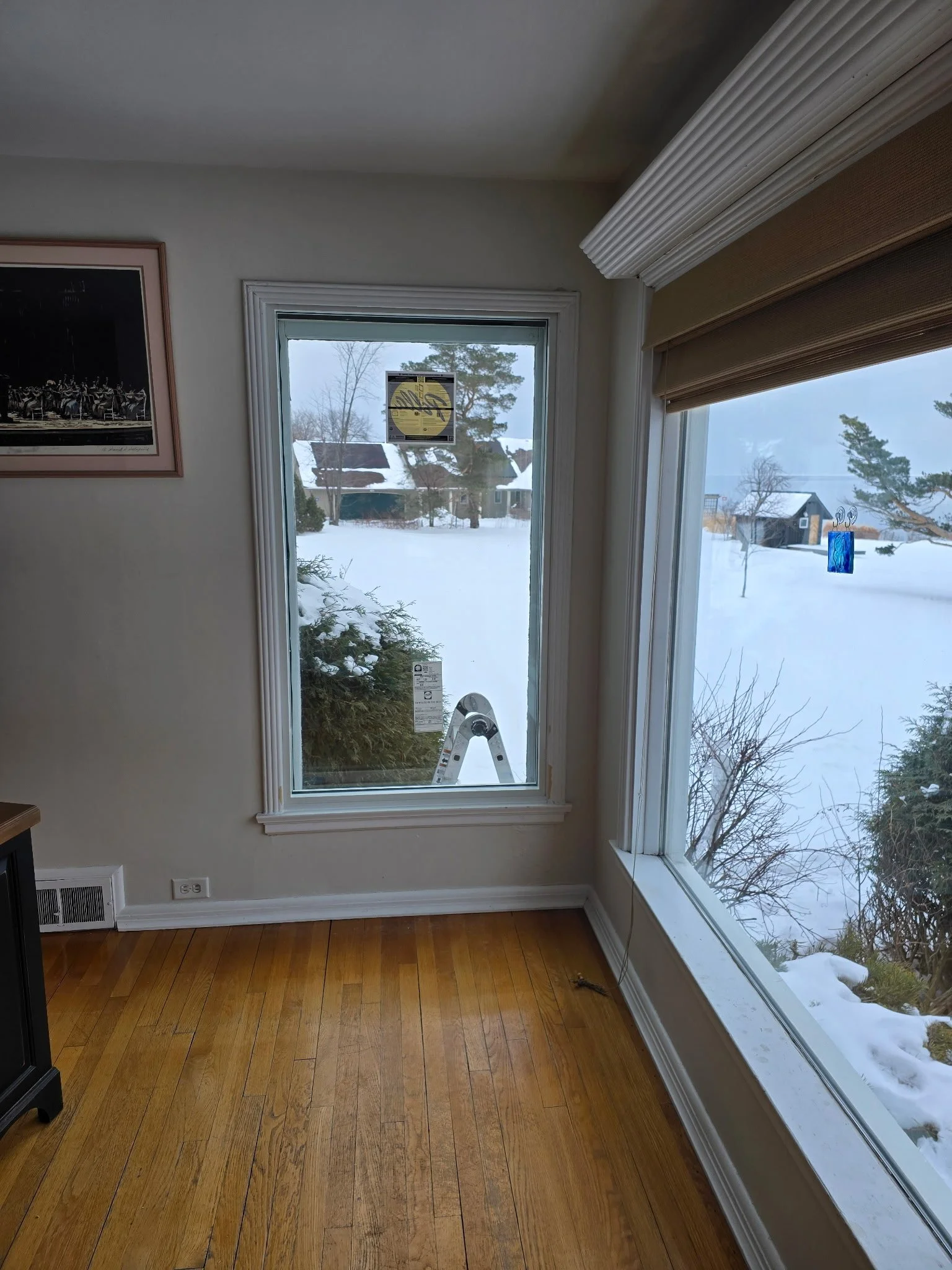 View of snow-covered yard and houses through large windows inside a room with wooden flooring.