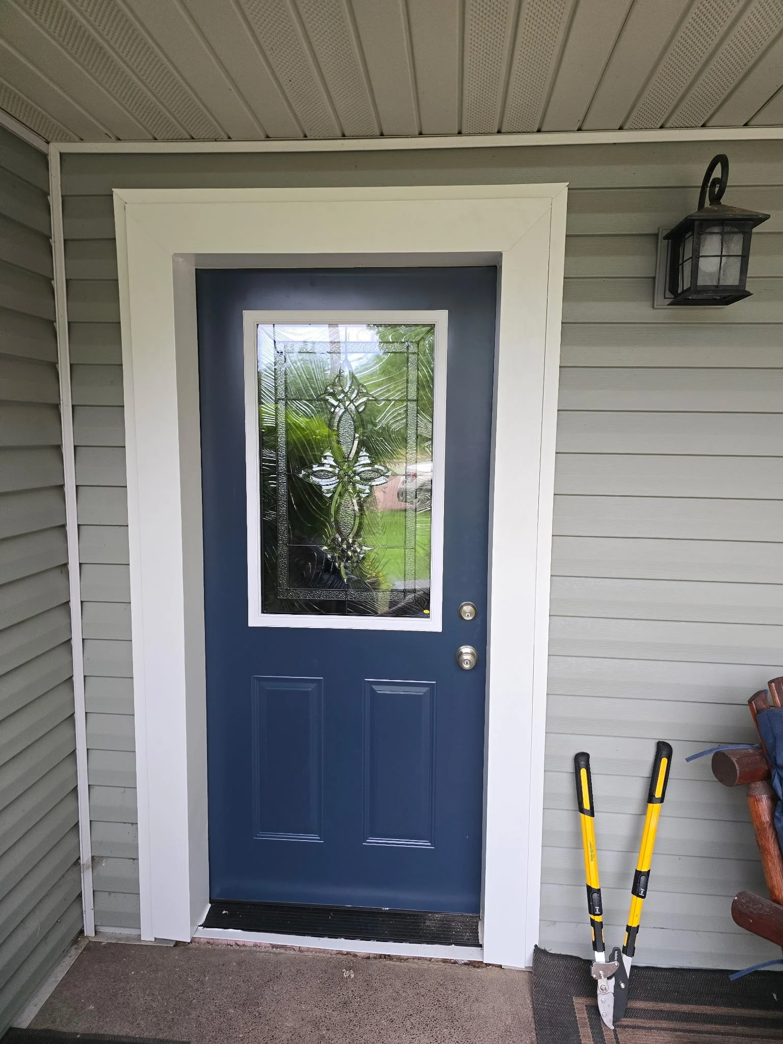 Blue front door with decorative glass window, white framing, and two door handles on a porch with grey siding and black outdoor lantern.