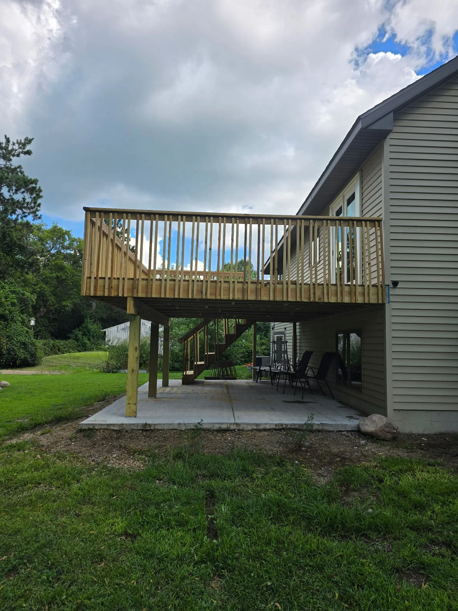 A newly built wooden deck attached to the back of a house, with a concrete patio below and outdoor chairs. The deck is elevated with vertical wooden railings and accessed by a staircase, surrounded by a yard with grass and trees.