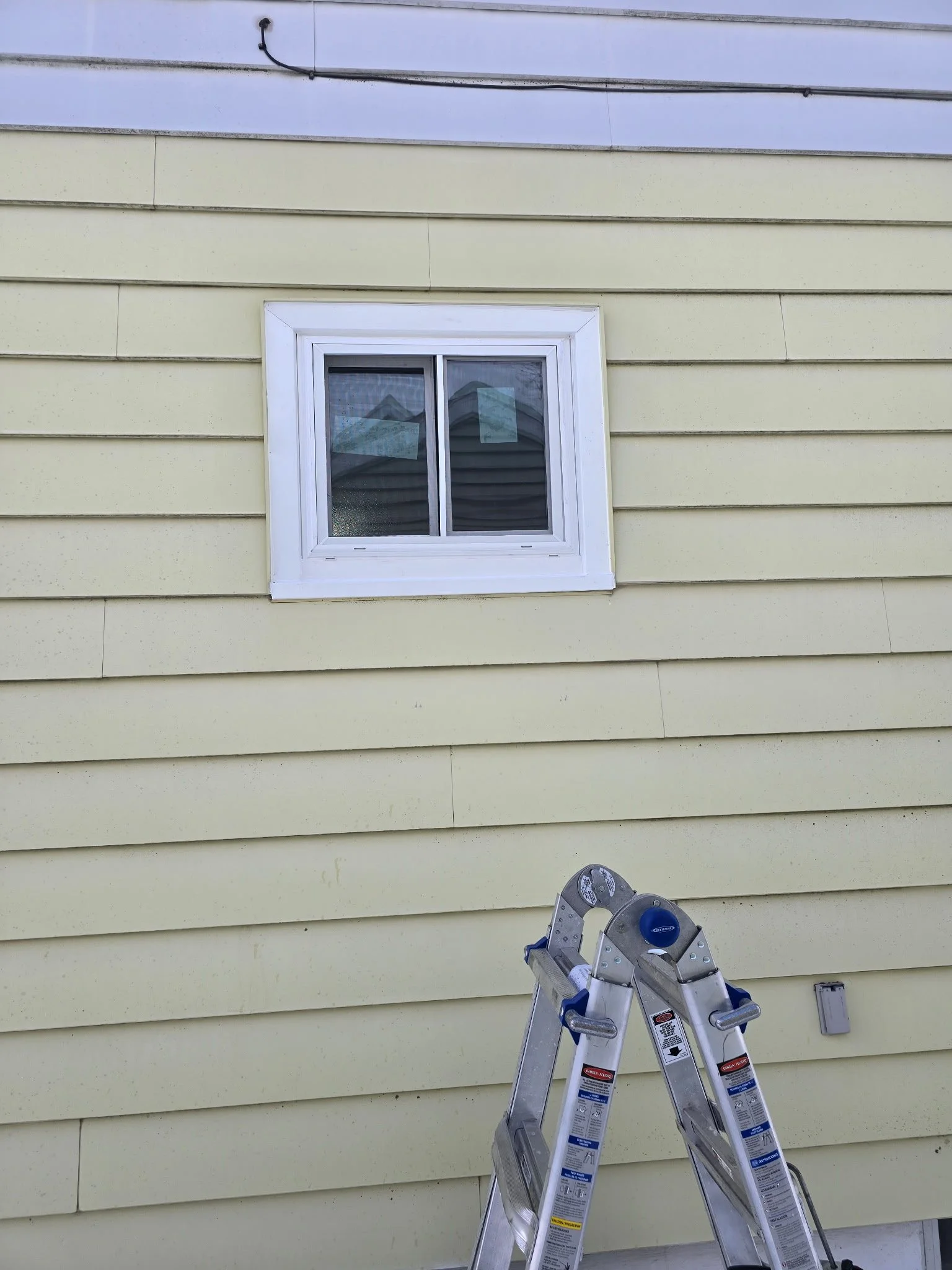 Exterior of a yellow house with a white-framed window, a ladder leaning against the wall, and a small outdoor electrical box.