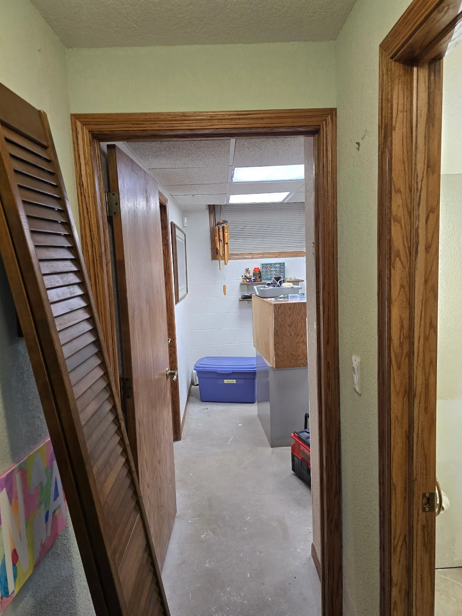 View of a basement or utility room with a concrete floor, wooden door frames, and a drop ceiling with fluorescent lights. There are shelves, storage boxes, and various tools visible.