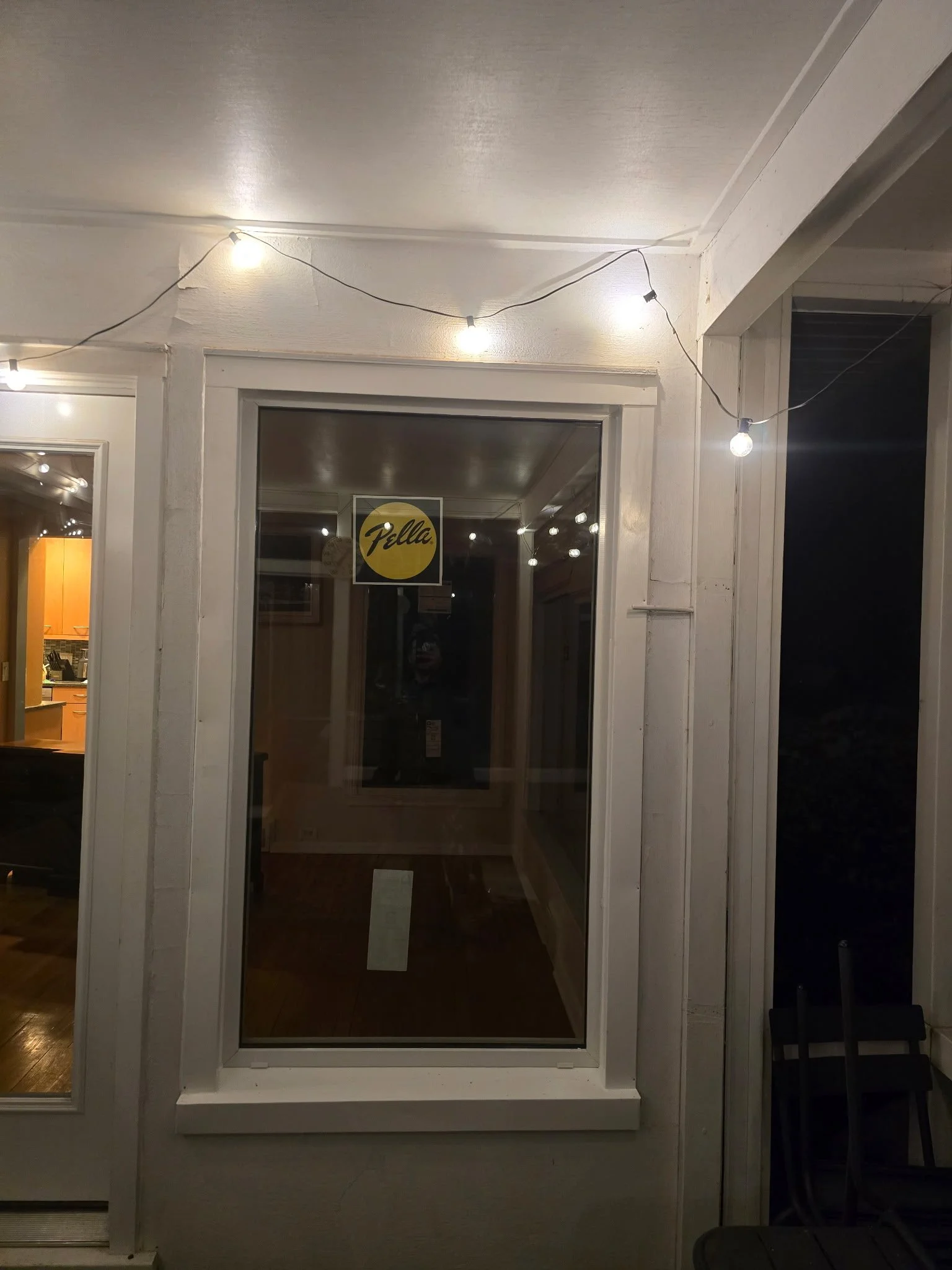 Nighttime view of a window with a yellow circle sticker labeled "Pella." String lights hang across the porch ceiling, and through the window, a kitchen with wooden cabinets is visible.