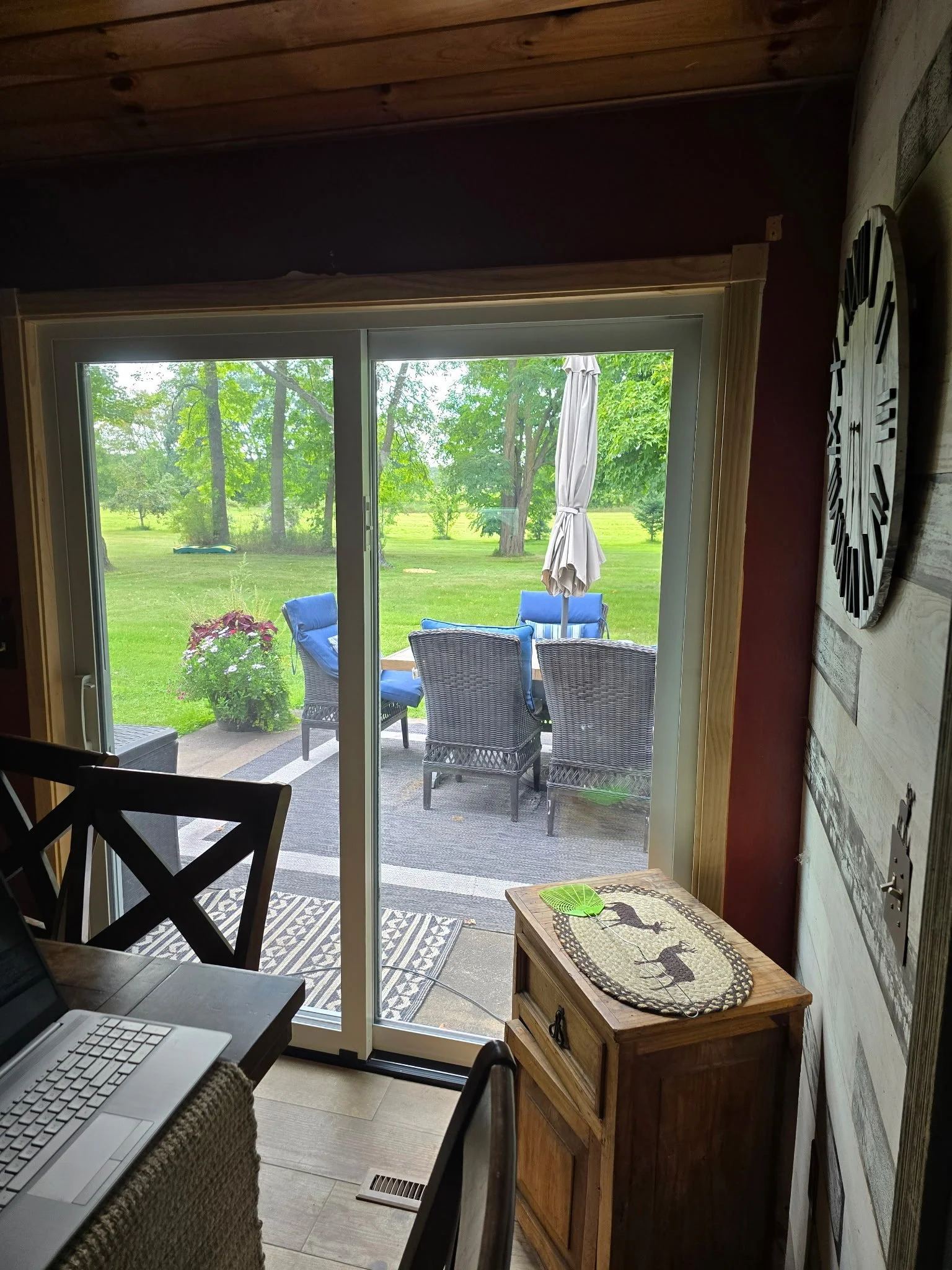 View from inside a house looking out through sliding glass doors onto an outdoor patio with chairs, a table with an umbrella, and green lawn with trees.