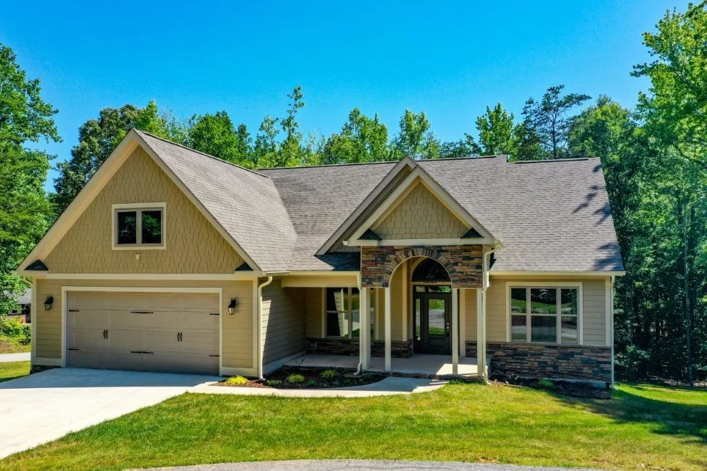 Front view of a modern house with a beige exterior, stone accents, a two-car garage, and a landscaped lawn surrounded by trees under a bright blue sky.