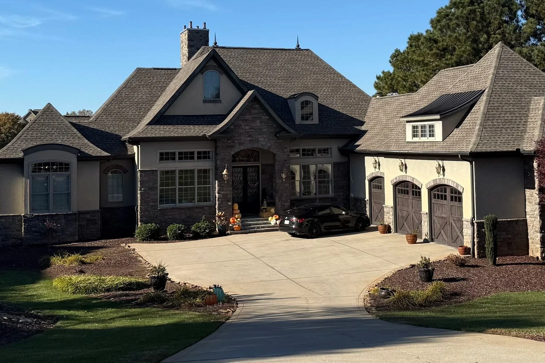 A large modern house with a multi-gable roof, stone accents, and a three-car garage, located on a landscaped lot with a curved driveway and decorative pumpkins in front.
