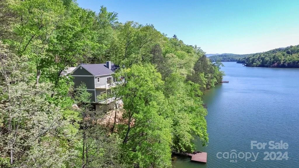 A house situated on a hillside surrounded by lush green trees, next to a large body of water on a clear, sunny day.