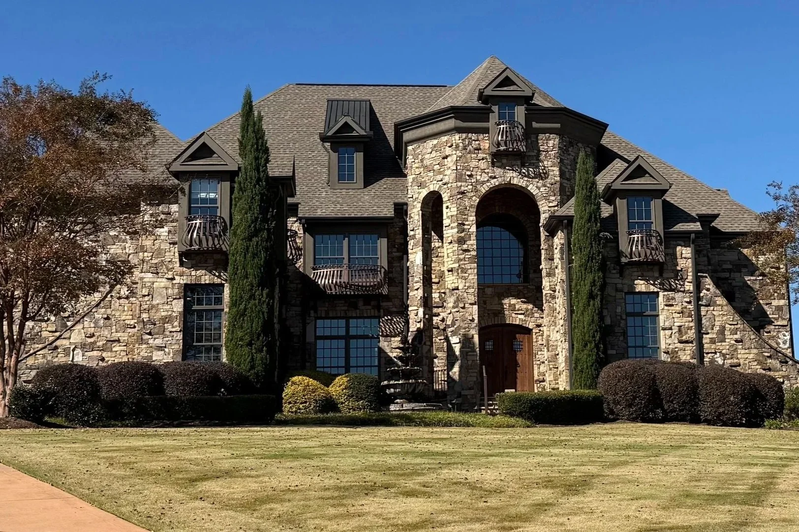 Large stone mansion with steep rooftop gables and small Juliet balconies, surrounded by well-kept bushes and grass, under a clear blue sky.