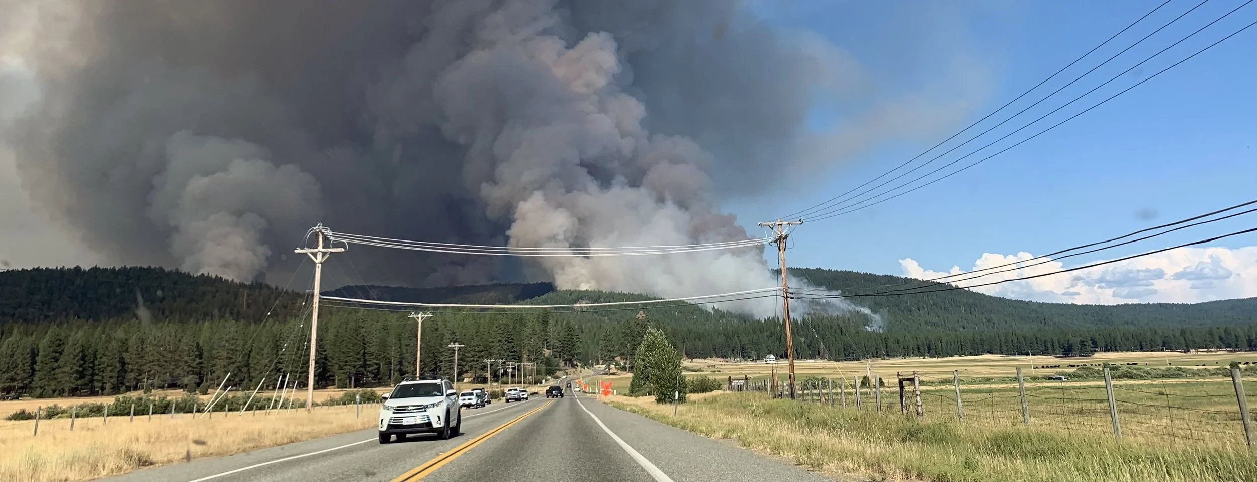Road with cars driving toward a wildfire in the distance, large black smoke billowing into the sky behind a forested hill.