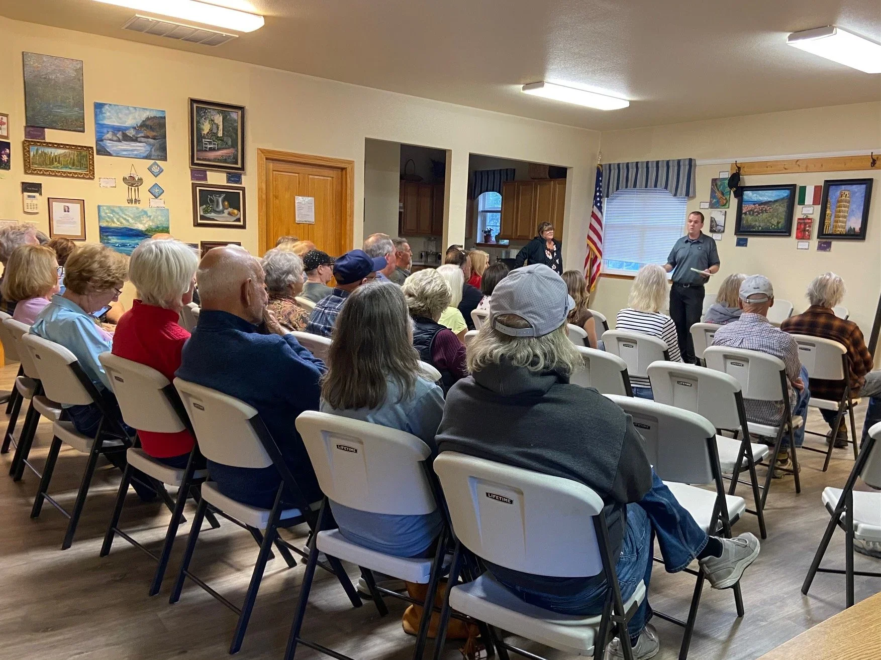 A group of people attending a presentation in a room with artwork on the walls, an American flag, and two presenters at the front.