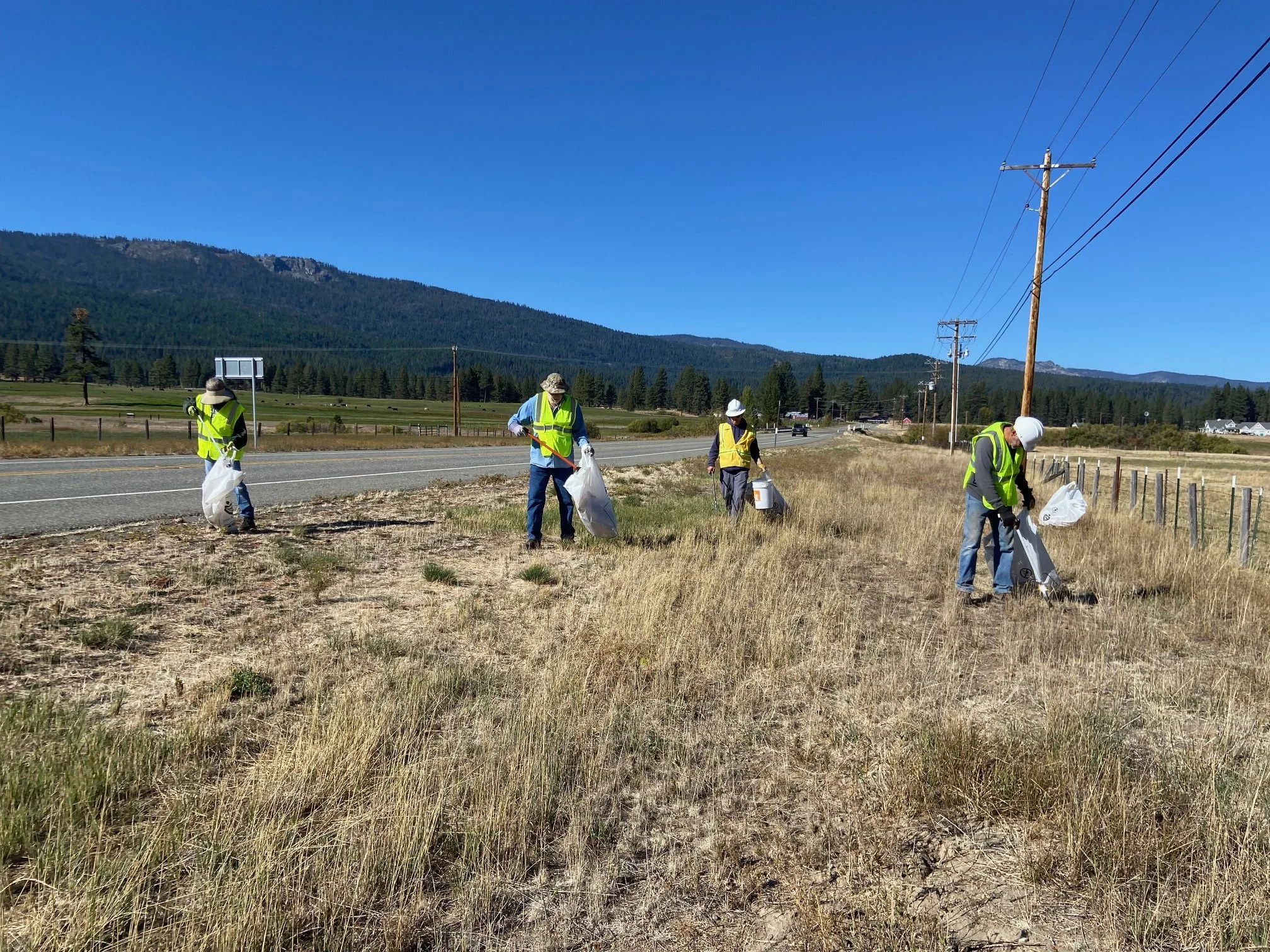 Four workers wearing safety vests and helmets picking up litter along a grassy roadside area with utility poles and mountains in the background.