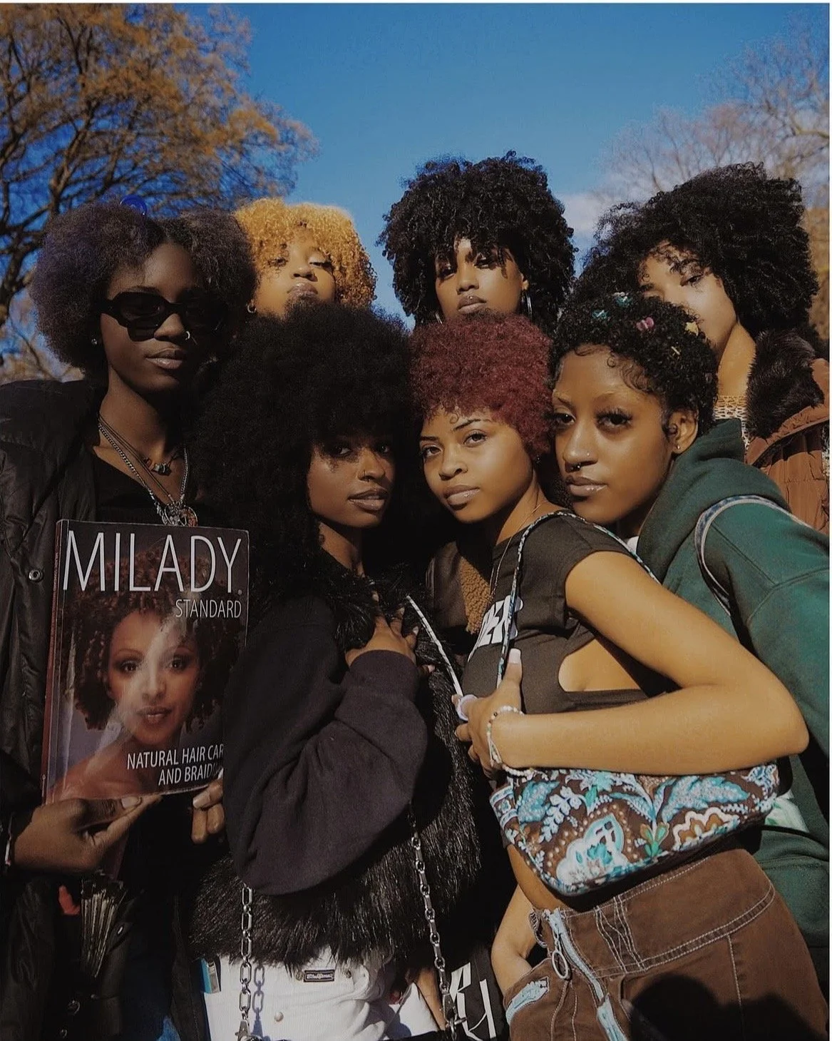 A group of eight women with diverse natural hairstyles standing outdoors against a clear blue sky and autumn trees in the background.