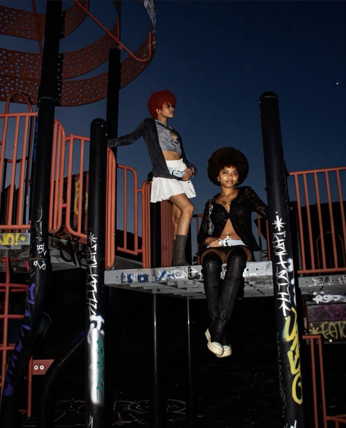 Two women with natural curly hair on a graffiti-covered playground structure during dusk. One is sitting, wearing a black lace jacket, black thigh-high boots, and a black and white outfit. The other is standing, wearing a dark jacket, grey crop top, white skirt, and black boots.