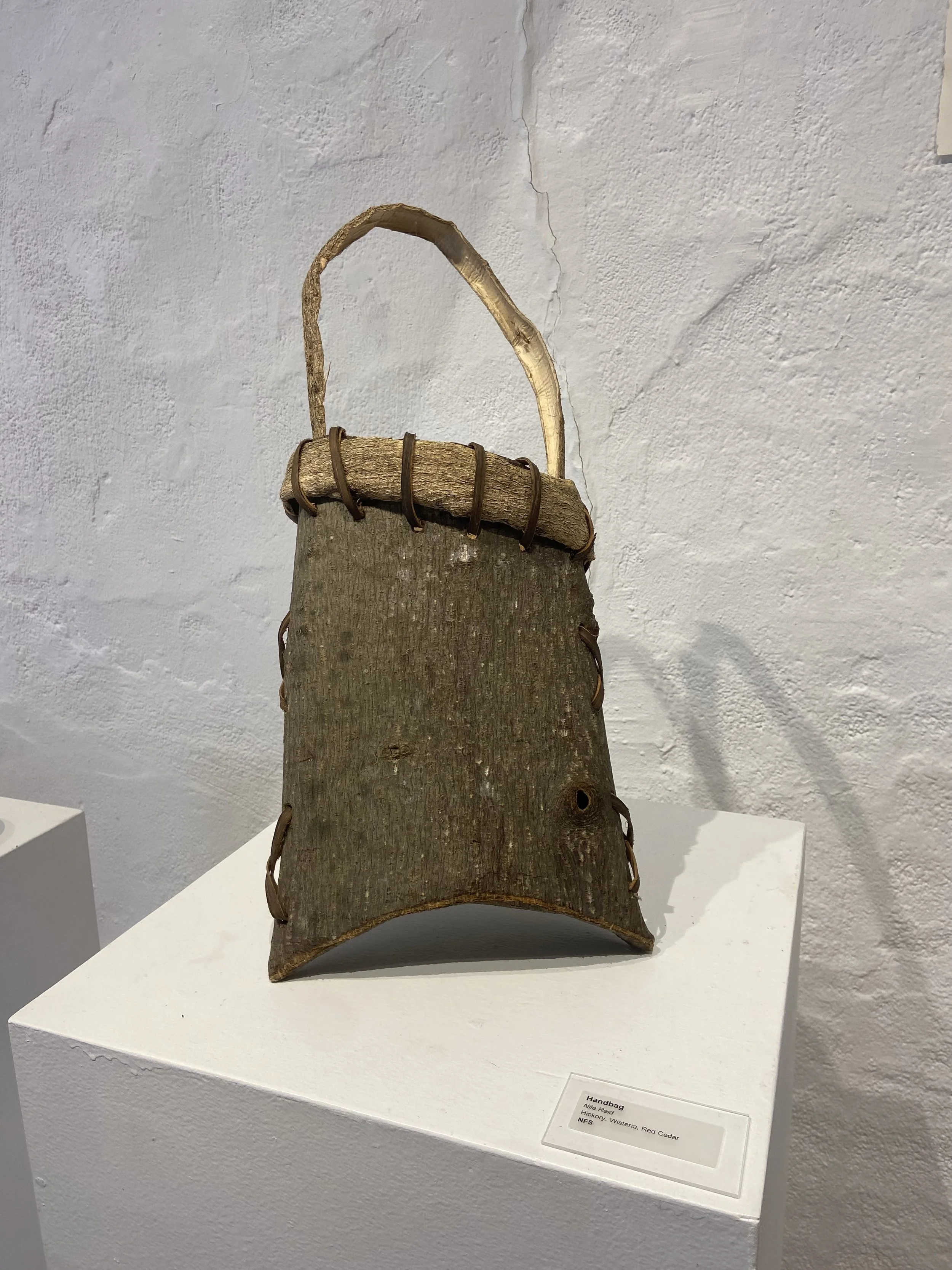 A rustic handcrafted bag made of wood and leather with a curved base, a handle, and leather straps along the sides, displayed on a white pedestal.