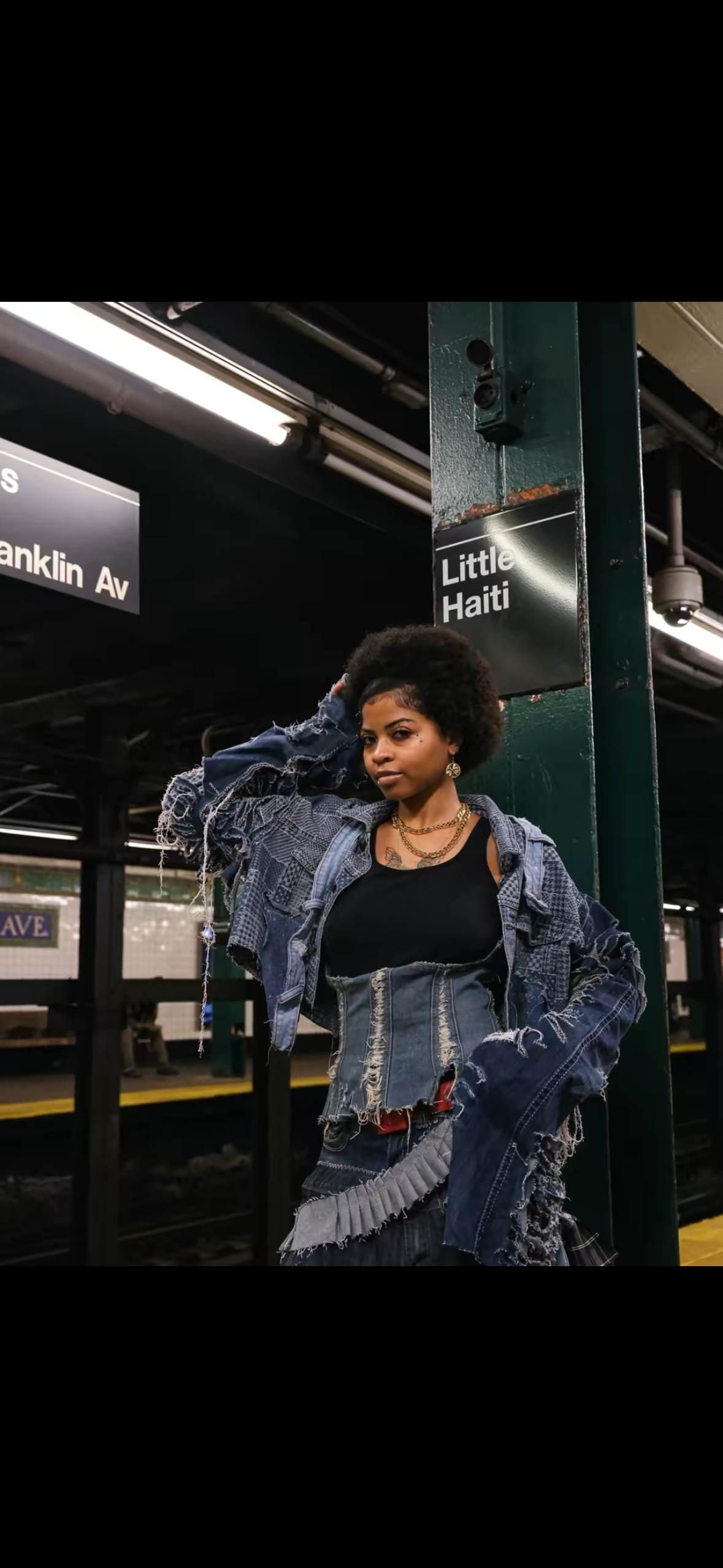A woman posing at a subway station on the Little Haiti platform, wearing a distressed denim jacket and a corset-style top, with her hand on her head.