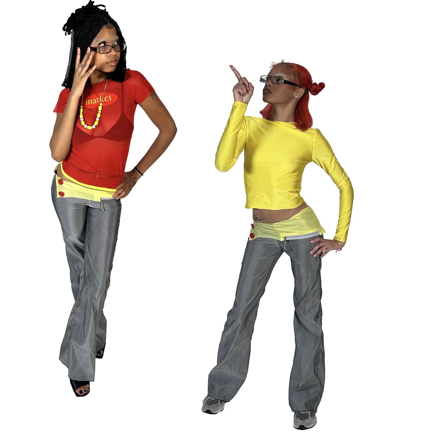Two women with colored hair and glasses posing against a white background. The woman on the left has dreadlocks, is wearing a red top with the word 'market' on it, and gray pants. The woman on the right has red hair in buns, is wearing a yellow long-sleeve crop top, and gray pants.