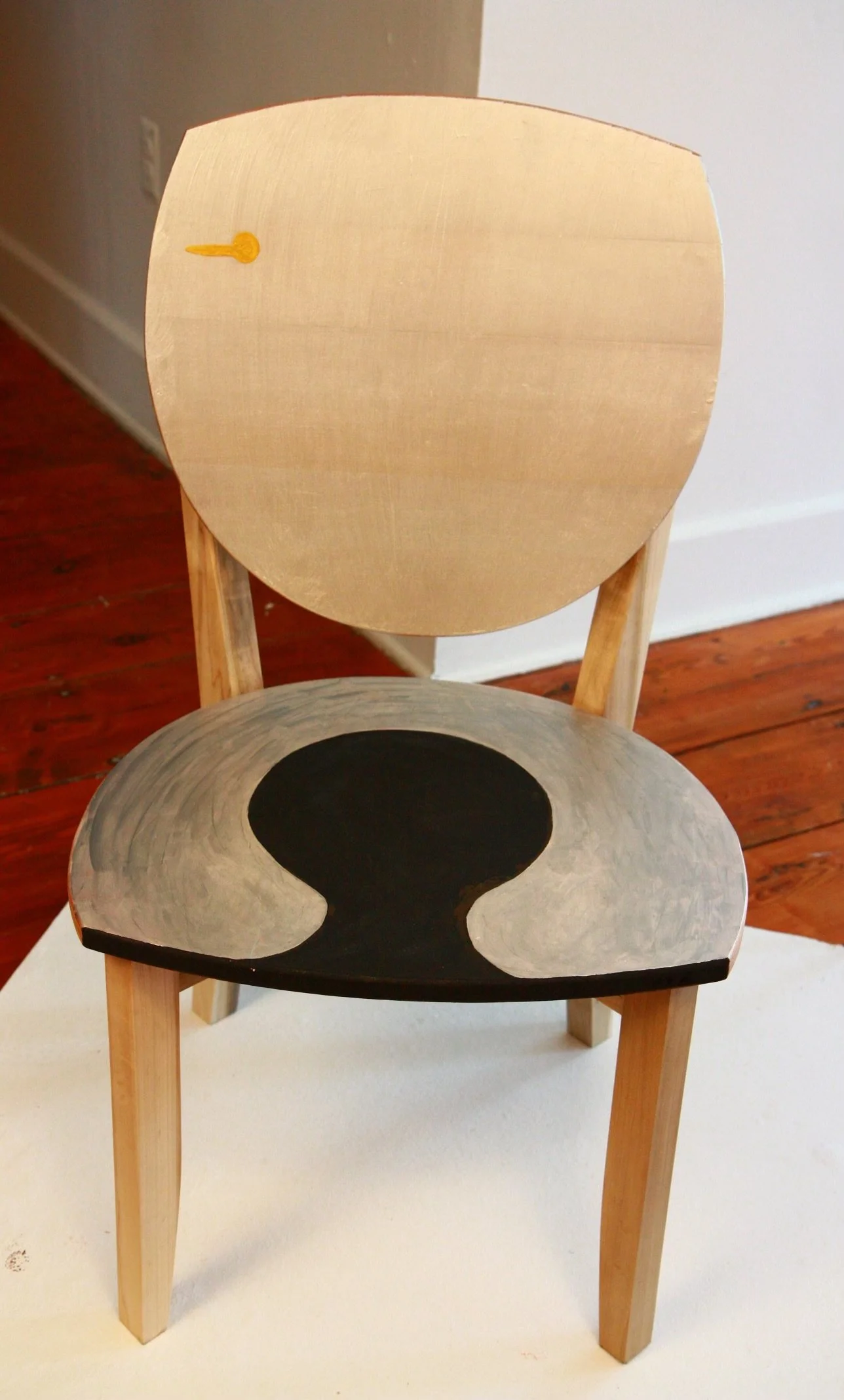 A wooden chair with a painted black and gray abstract design on the seat and a plain wooden backrest, situated on a white surface with a background of a beige wall and wooden floor.