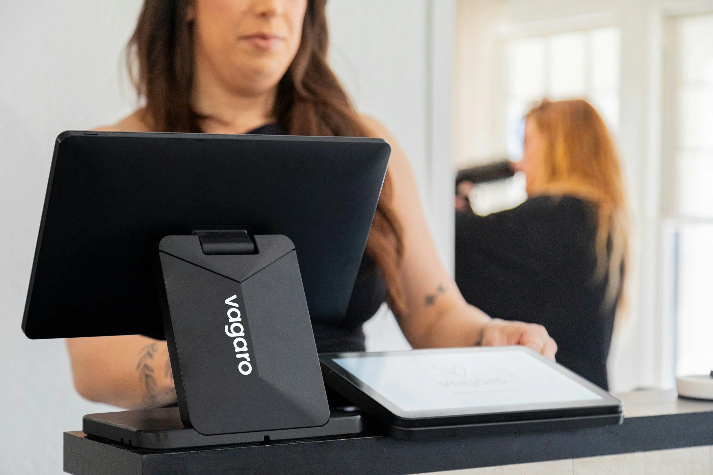 A woman using a Verifone mobile payment terminal at a counter, with another woman in the background drinking coffee.