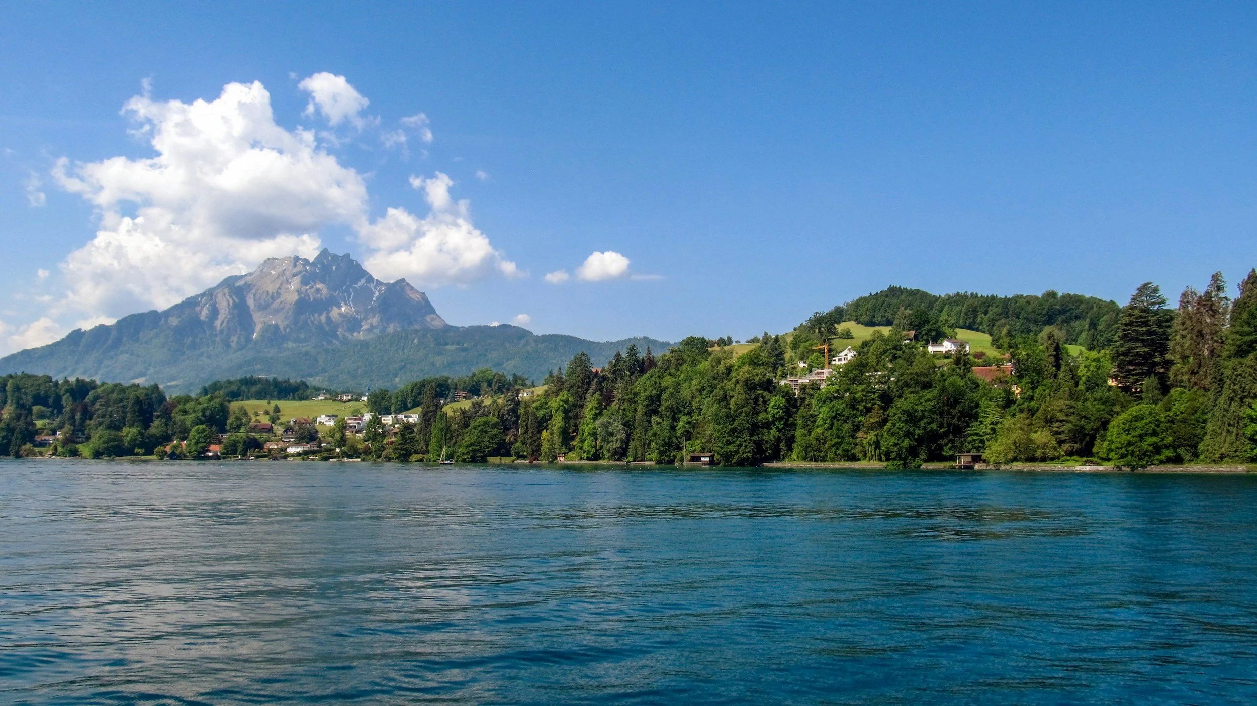 Scenic view of a mountain with lush green trees and houses by a large lake under a bright blue sky with white clouds.