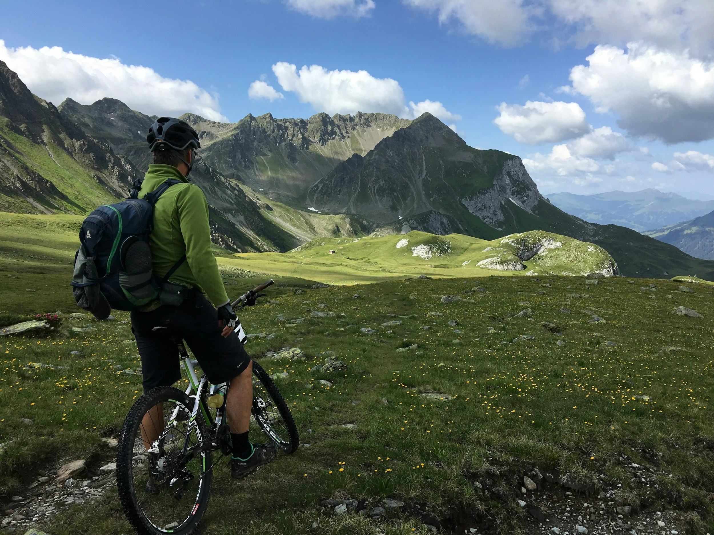 A person with a backpack and helmet riding a mountain bike in a lush green valley surrounded by mountains under a partly cloudy sky.