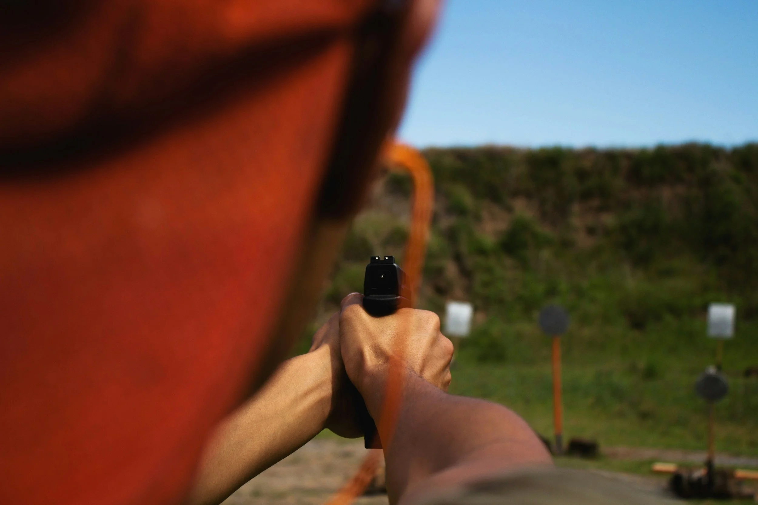 Person firing a handgun at outdoor shooting range, with target boards in distance and hills in background.