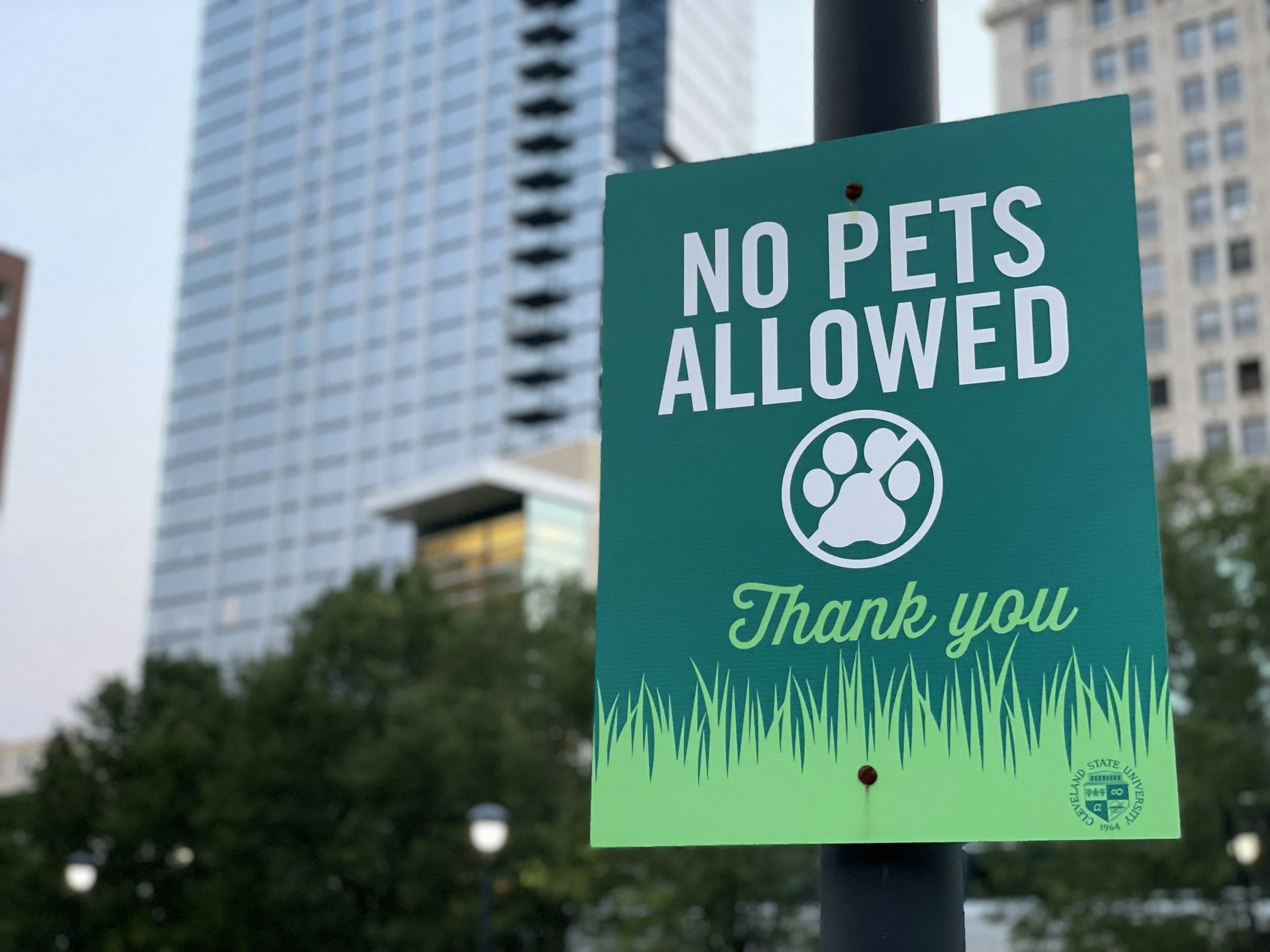 Sign that says 'No Pets Allowed' with a paw print symbol and a 'Thank you' note at the bottom, located outdoors with trees and tall buildings in the background.