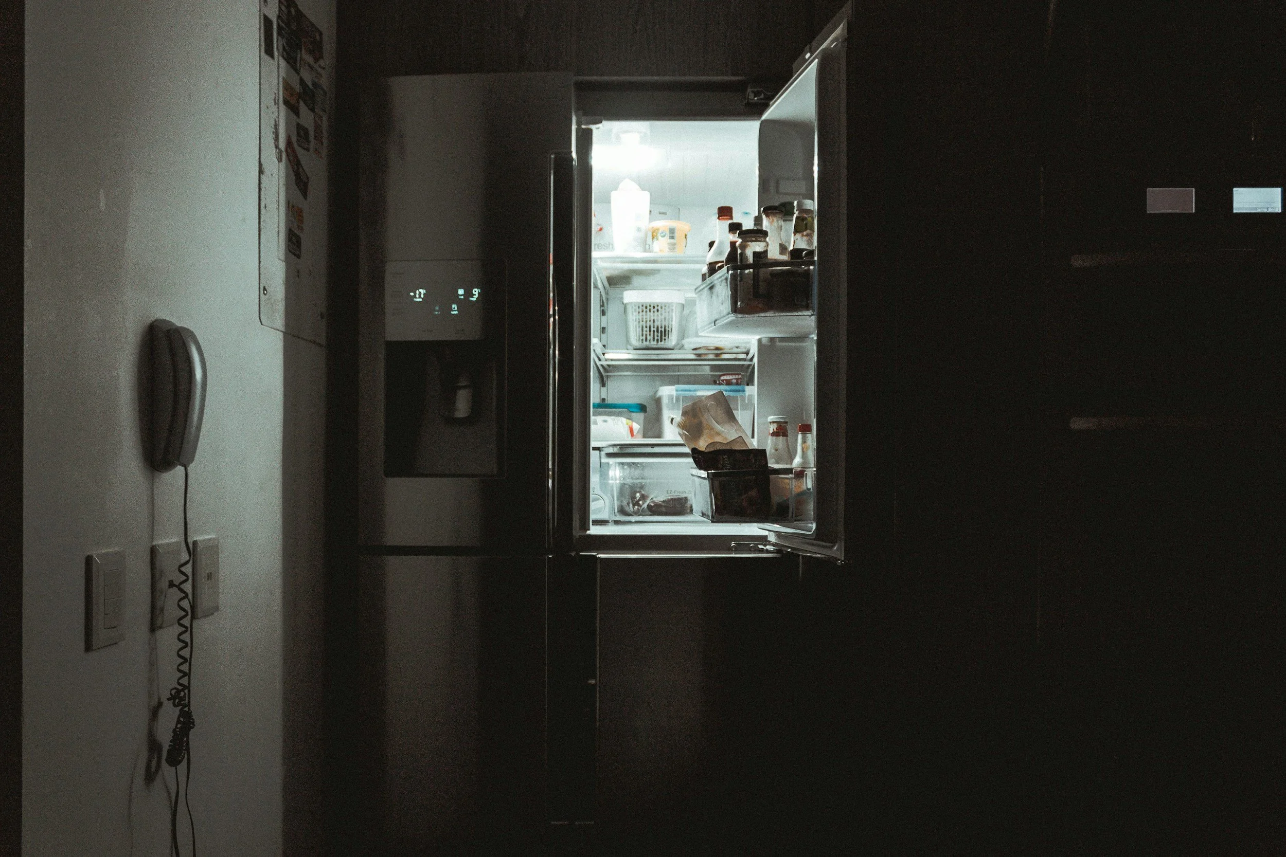 Open refrigerator with various food and beverage items inside, illuminated amidst a dark room, with a wall-mounted telephone and switches to the left.