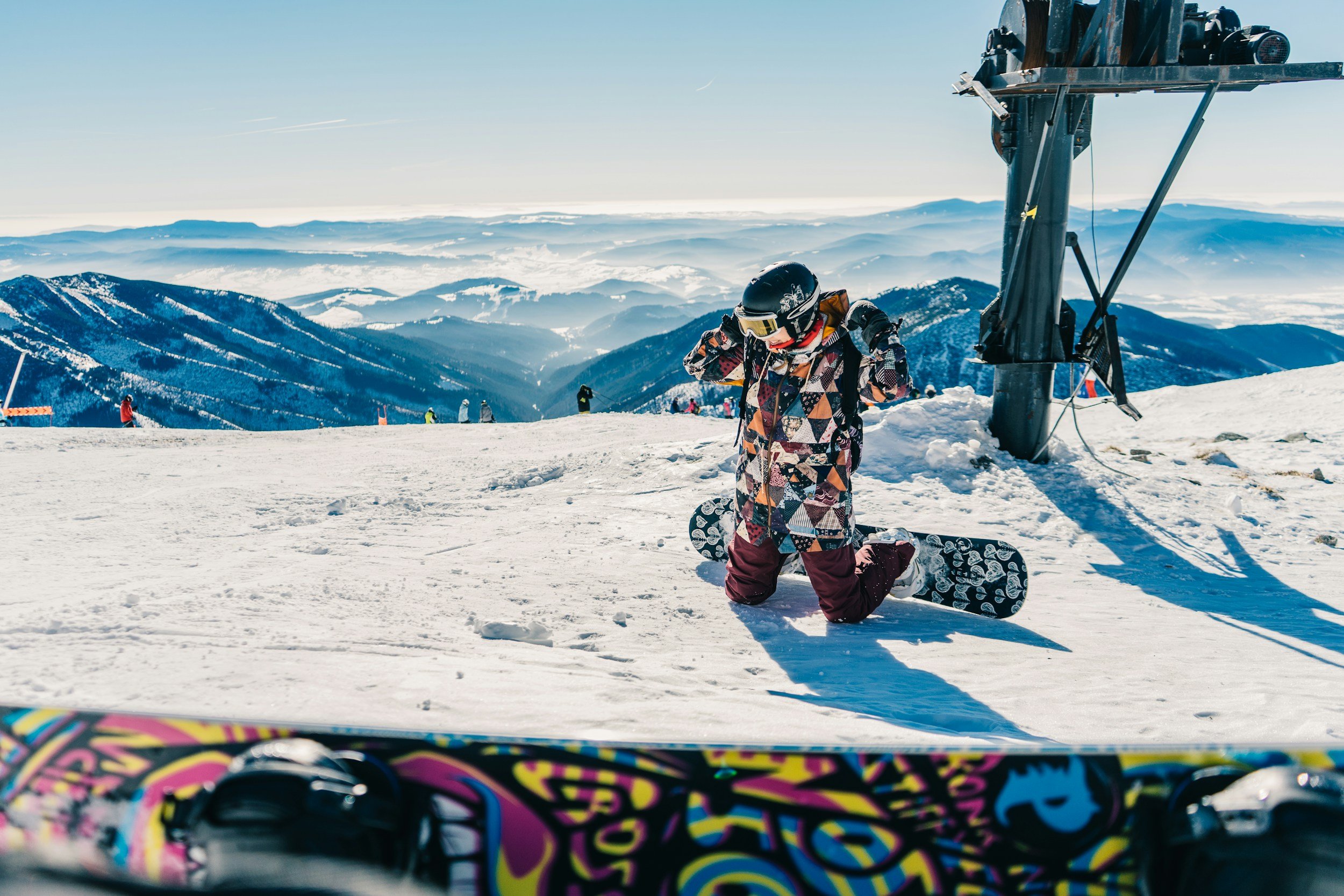 Child wearing a snowboard helmet and goggles adjusting gear on a snowy mountain slope with snow-covered mountains and a clear sky in the background.