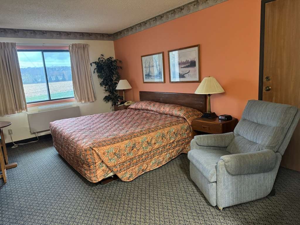A motel room with a bed, two nightstands with lamps, a grey armchair, and a window with curtains showing a view of trees and a field