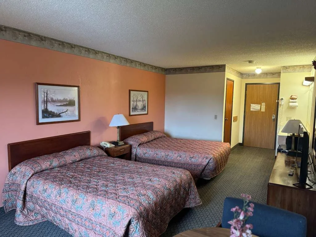 A hotel room with two queen beds featuring patterned bedding, a wooden headboard, and a nightstand with a lamp and telephone. The room has a pinkish-orange accent wall with framed landscape paintings, a television on a dresser, and a small dining tab
