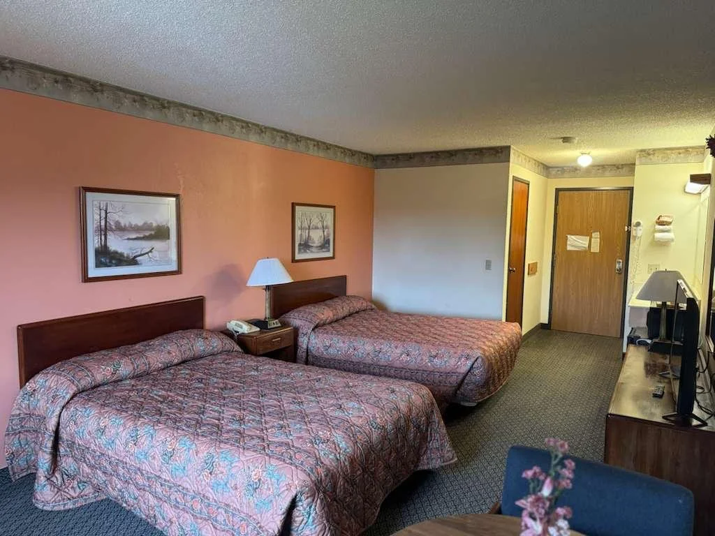 Hotel room with two double beds, a nightstand with a lamp and phone between them, paintings on the wall, and a flat-screen TV on a cabinet opposite the beds.
