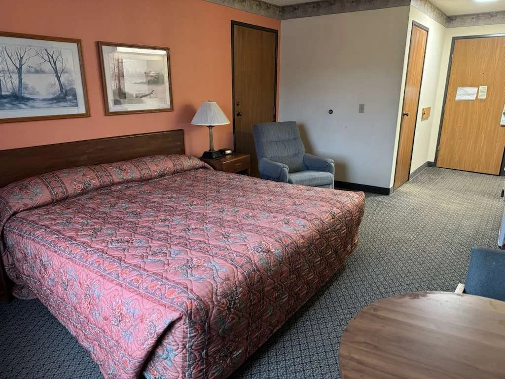 Hotel room with a large bed covered in a patterned red quilt, a wooden headboard, two paintings of winter scenes, a bedside lamp, and a blue armchair near the door.