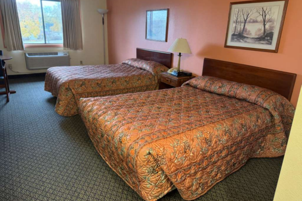 Two double beds with patterned bedspreads in a hotel room, with a nightstand, lamp, wall art, window, and air conditioning unit.