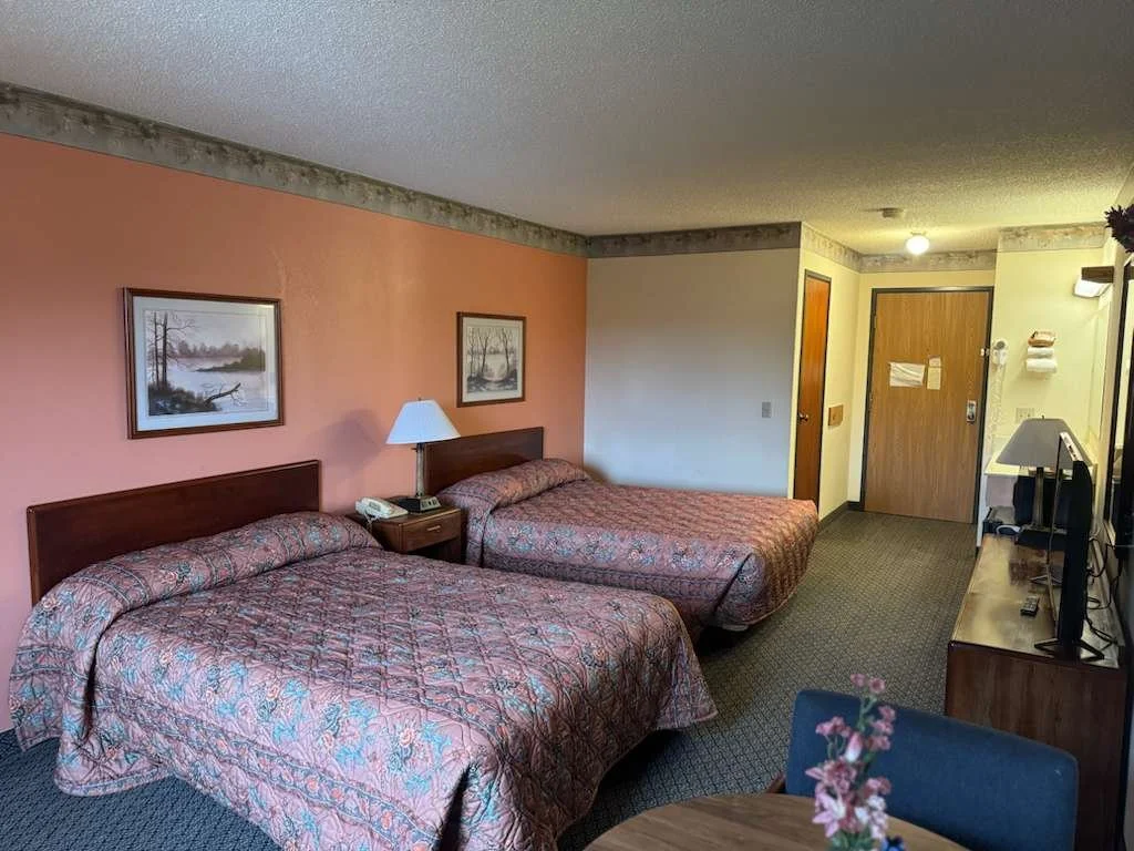 A hotel room with two double beds, a shared nightstand with a lamp and phone, a framed picture above each bed, a TV on a wooden dresser, and an entrance door in the background.