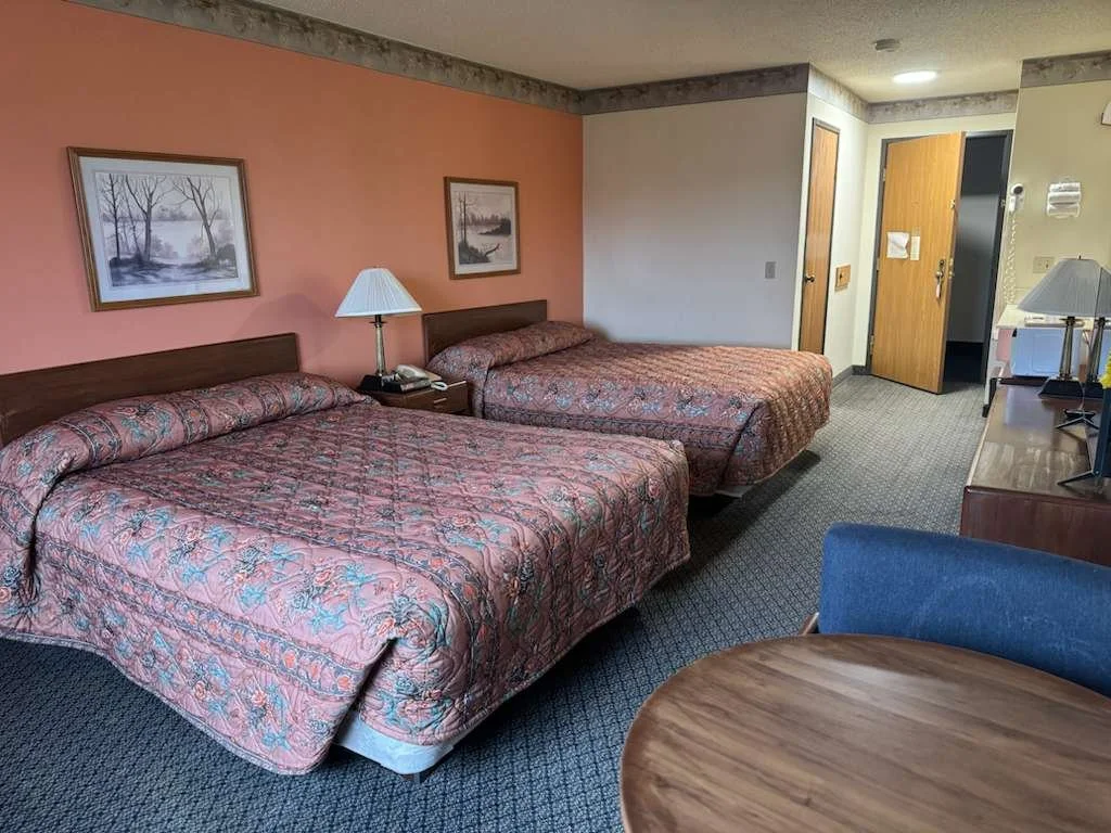 Hotel room with two double beds, a nightstand, two lamps, wall art, a desk with a lamp, and a door leading to the bathroom.