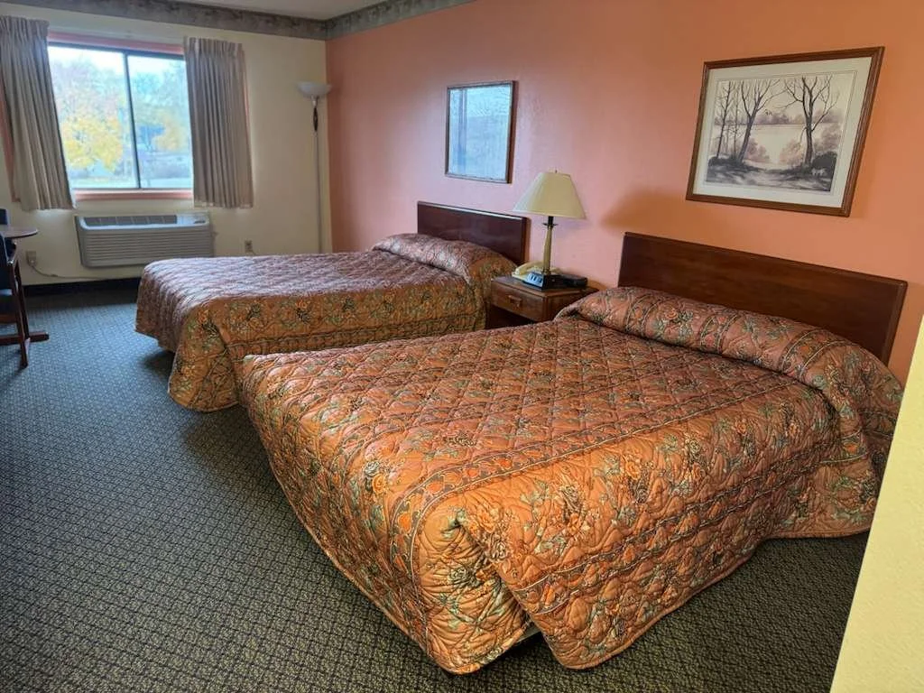 A motel room with two double beds covered in quilted bedspreads, a window with curtains, a nightstand with a lamp and phone, and framed pictures on the walls.