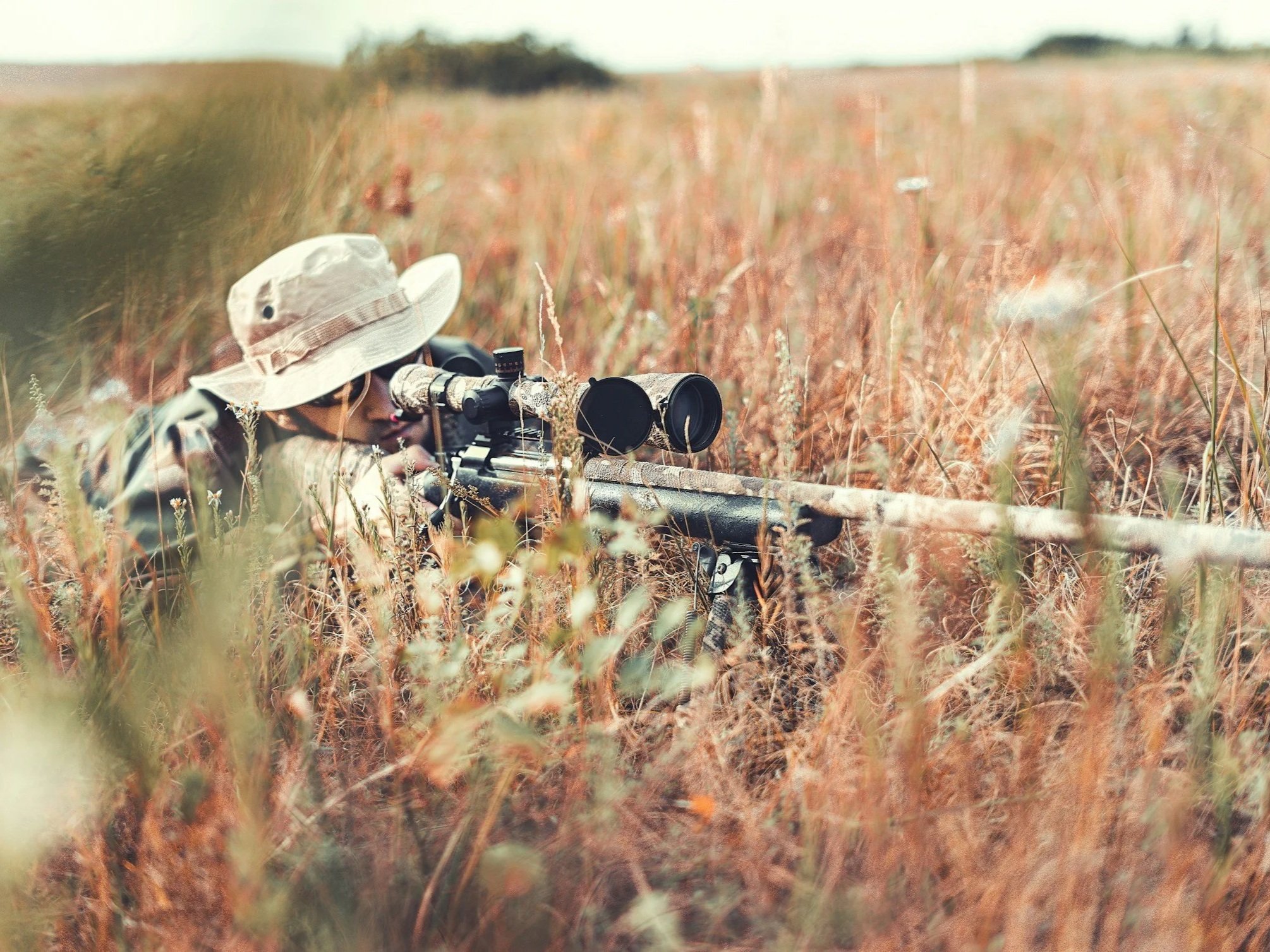 Person in camouflage clothing and wide-brimmed hat lying on the ground aiming a sniper rifle with scope in tall grassy field.