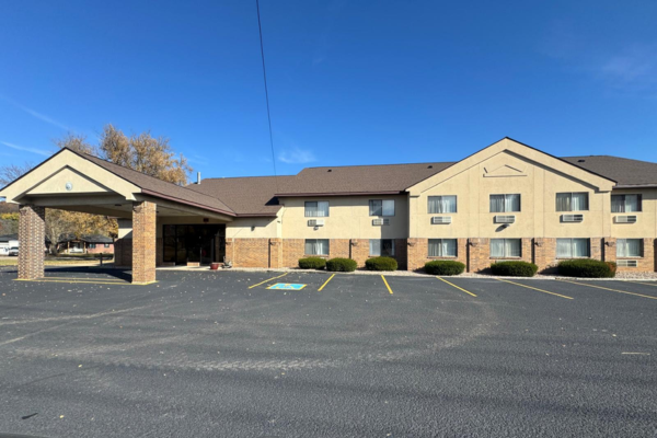 Exterior of a beige two-story motel or hotel building with brick accents, a covered entrance, and an empty parking lot with designated handicapped parking spots.