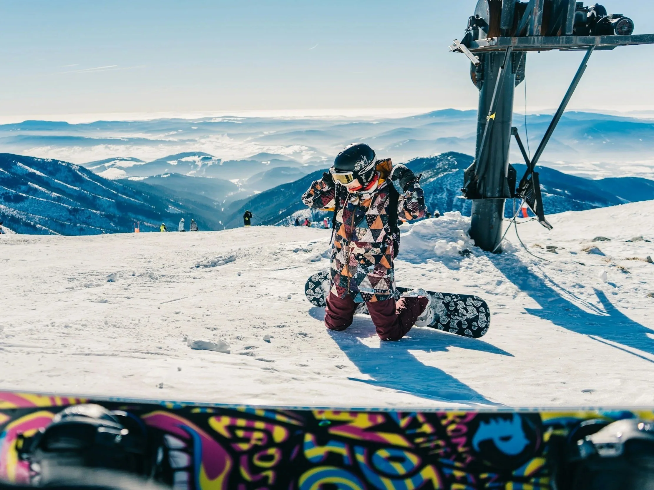 A child in winter gear kneeling on snow with a snowboard, mountain scenery in the background, near ski lift equipment on a bright sunny day.