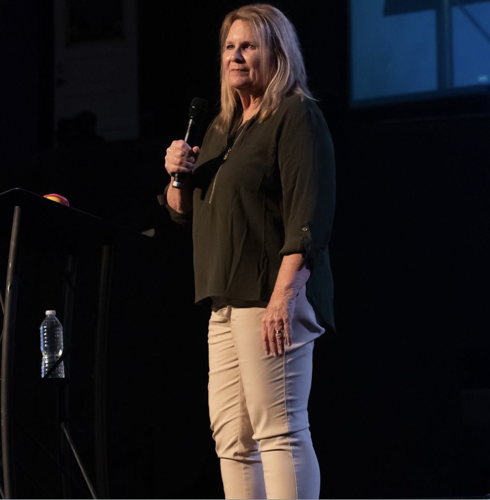 A woman with blonde hair stands on a stage, holding a microphone, wearing a dark green blouse and beige pants.